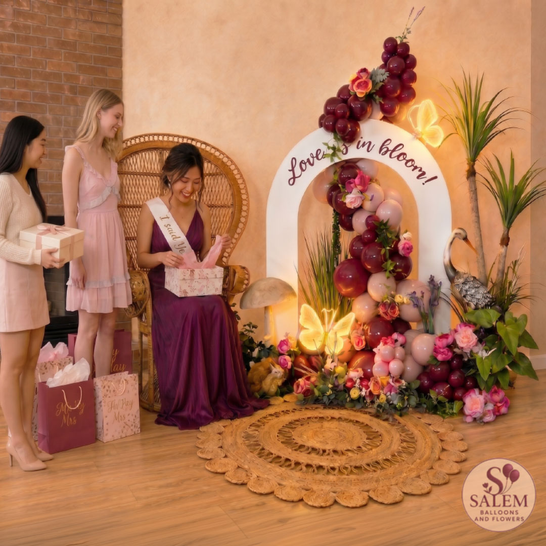 Woman seated with sash, surrounded by three girls, floral display with sign, and decorative elements in a room.