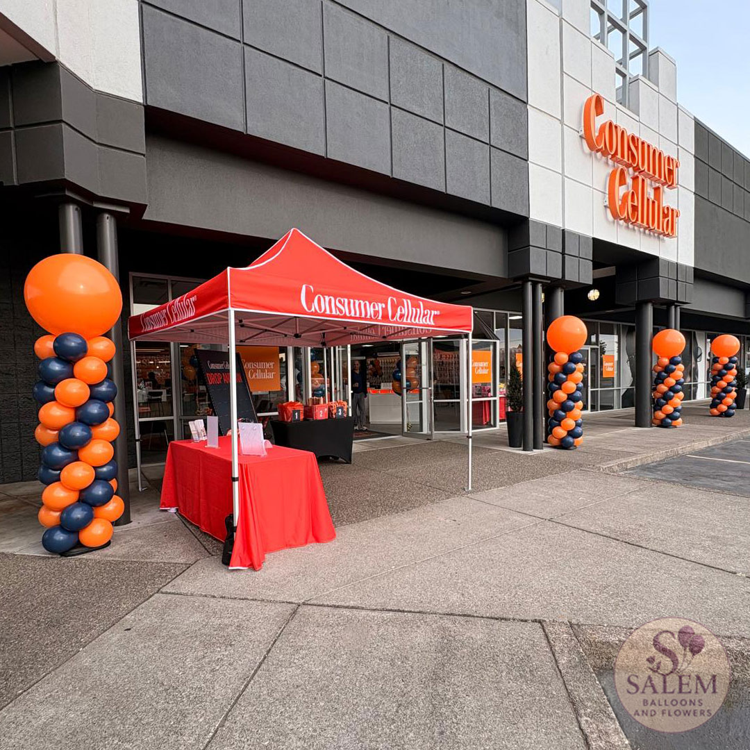 Red canopy tent with tables outside a store, decorated with orange and black balloon columns, in front of a shopping center.