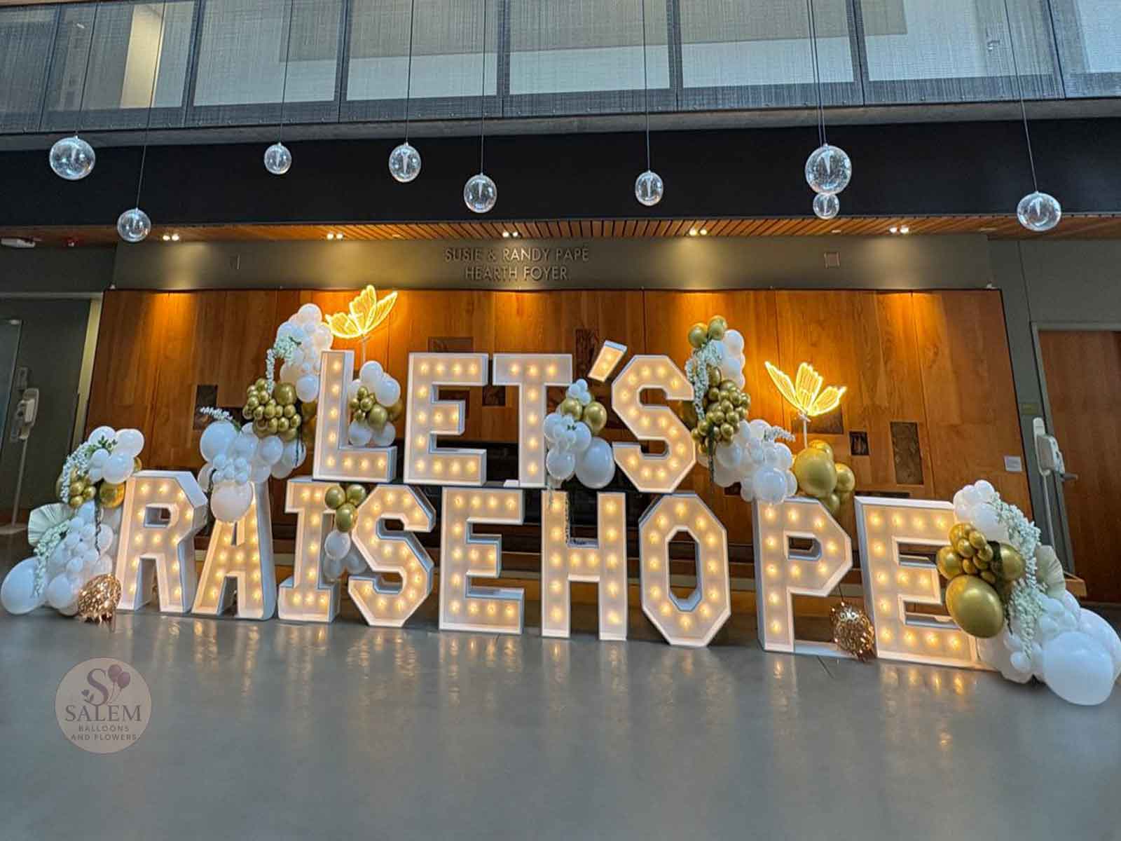 Decorative display with large illuminated letters spelling 'LET'S RAISE HOPE', surrounded by flowers, balloons, and hanging ornaments.