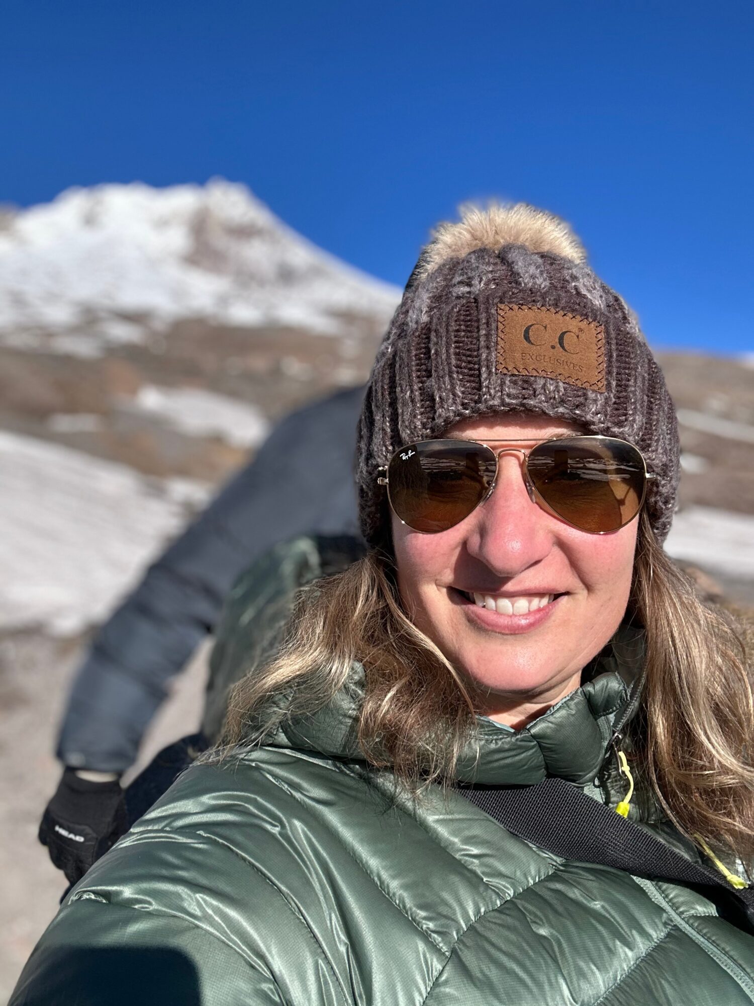Woman outdoors wearing sunglasses, a knit hat, and a green jacket, with snow-covered mountains in the background.