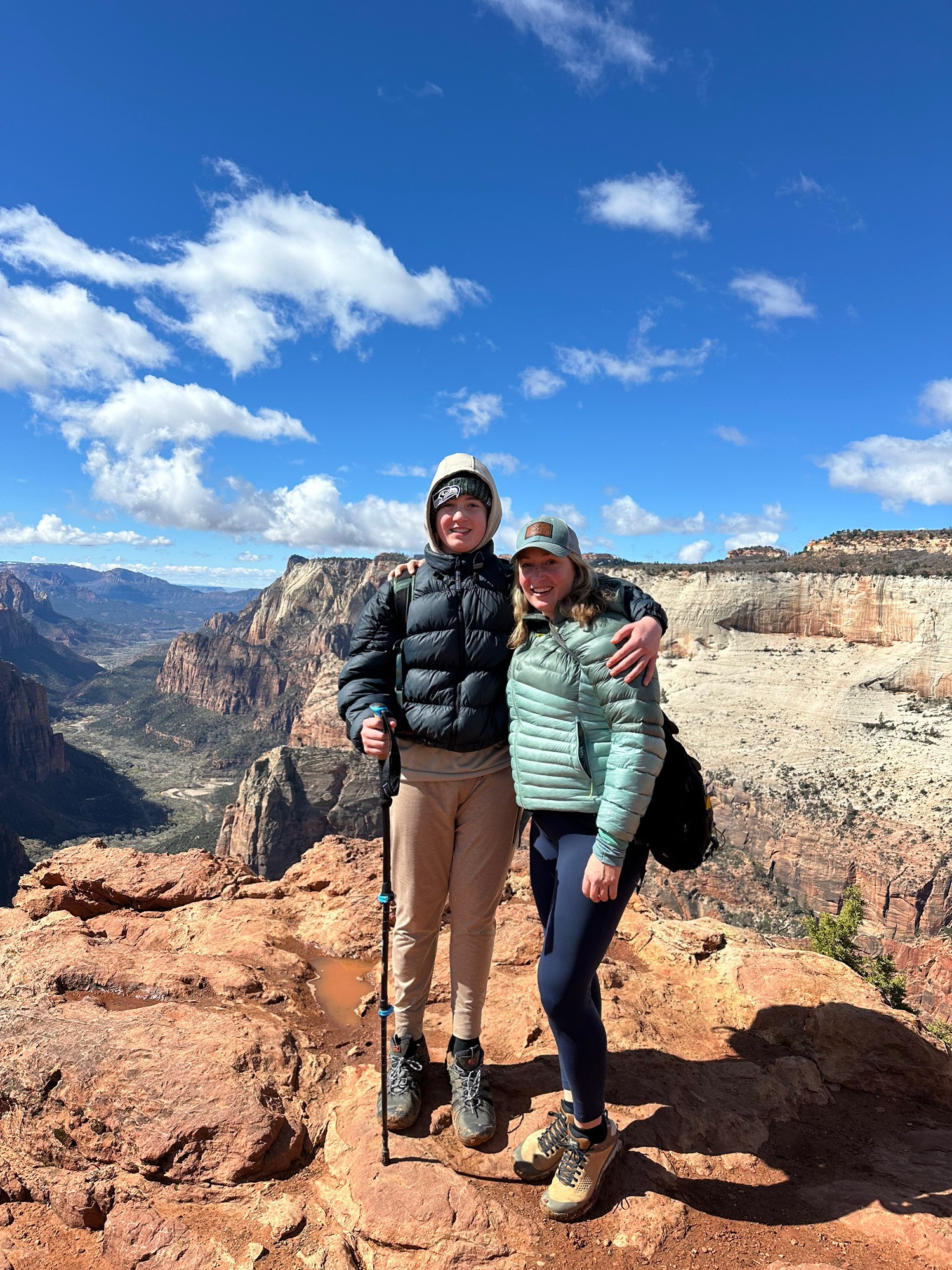 Two people stand on rocky terrain with a canyon and blue sky with clouds in the background, smiling and embracing.