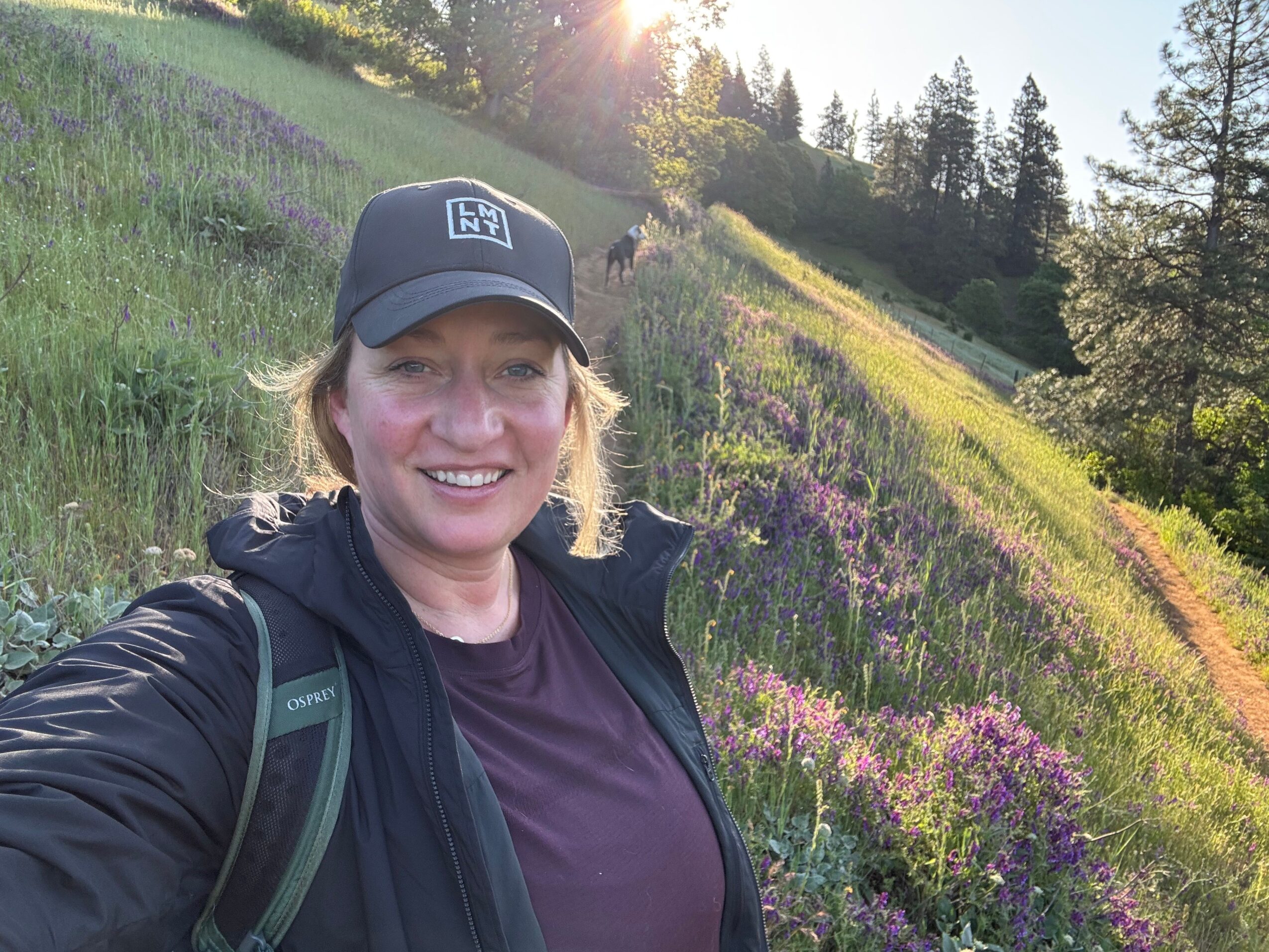 Person outdoors smiling, wearing a cap and backpack, with a landscape of trees and flowers in the background.
