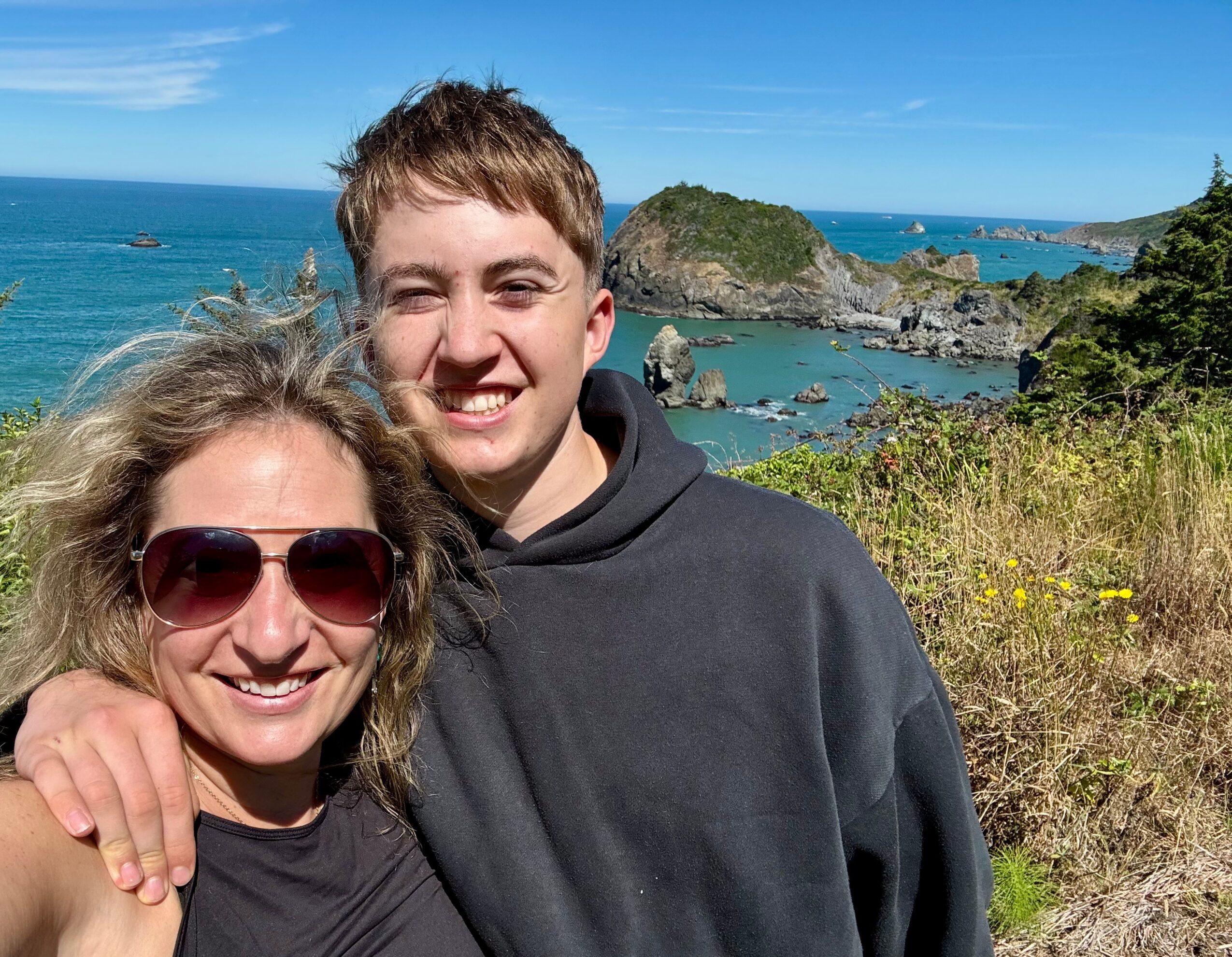 Two people smiling outdoors with a coastal landscape, rocks, and blue sky in the background.