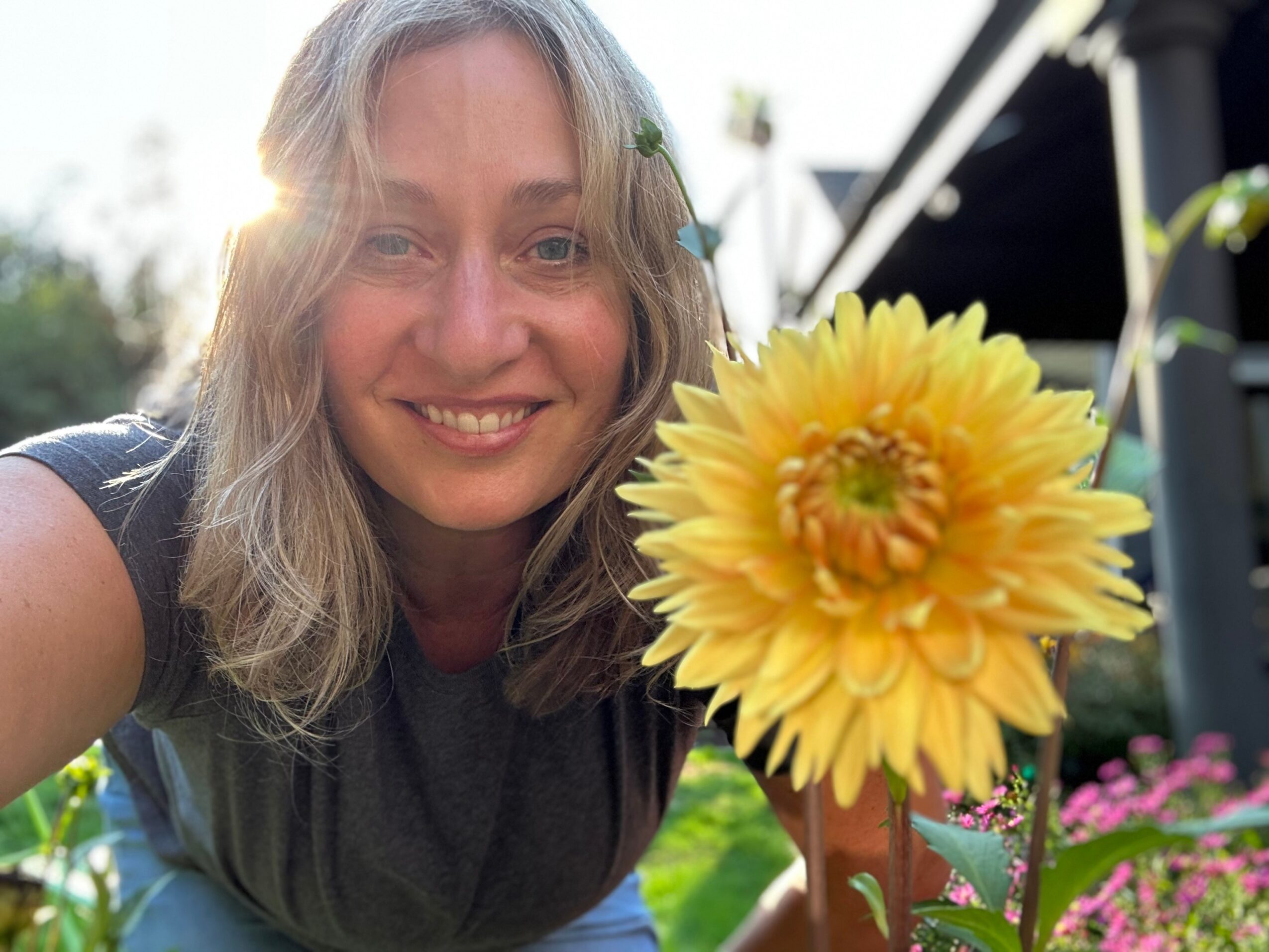 Woman smiling outdoors with a large yellow flower in foreground, sunlight behind her, greenery and flowers around.