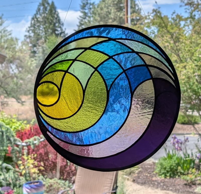 Colorful stained glass circular ornament with blue, green, yellow, and purple sections hanging outdoors with trees and flowers in background.