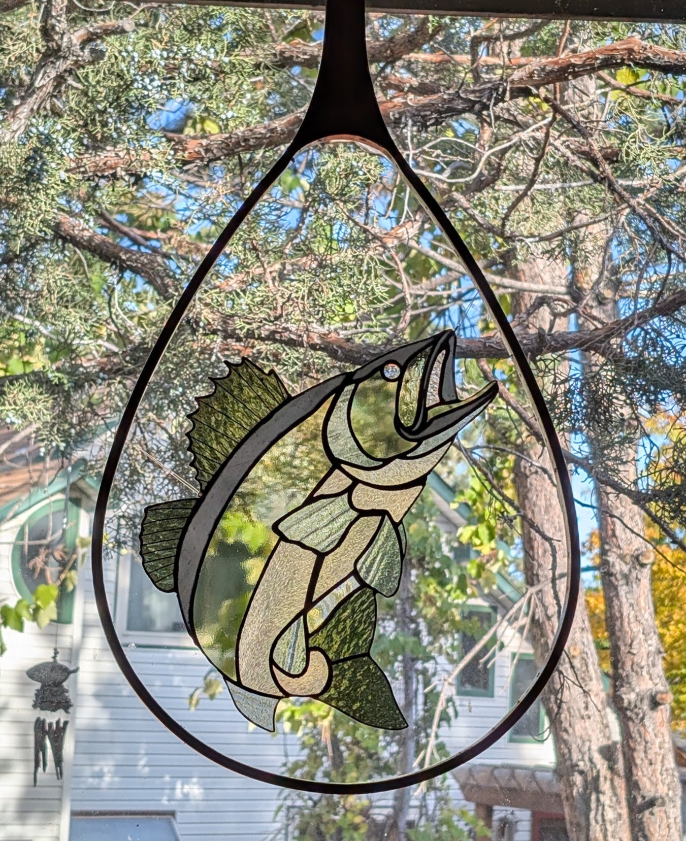 Decorative fish illustration inside a teardrop-shaped frame, hanging outdoors with trees and sky in background.