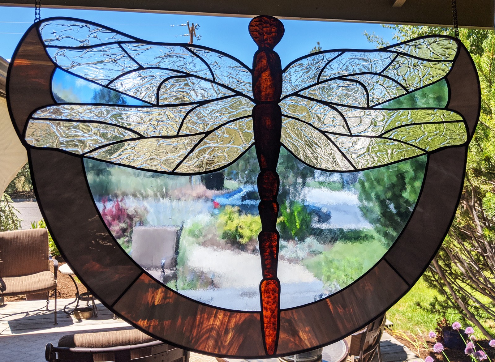 Colorful stained glass dragonfly with detailed wings and body, hanging indoors with a garden view outside.
