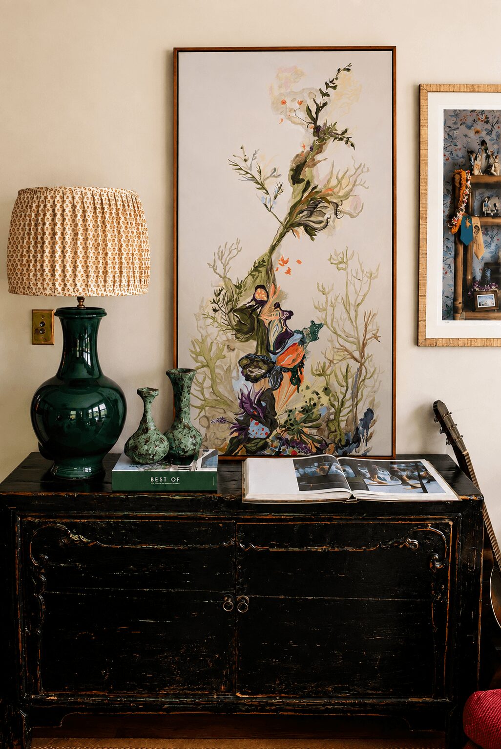 Decorative floral artwork with birds, a green ceramic lamp, and framed pictures on a wall, on a dark wooden cabinet.