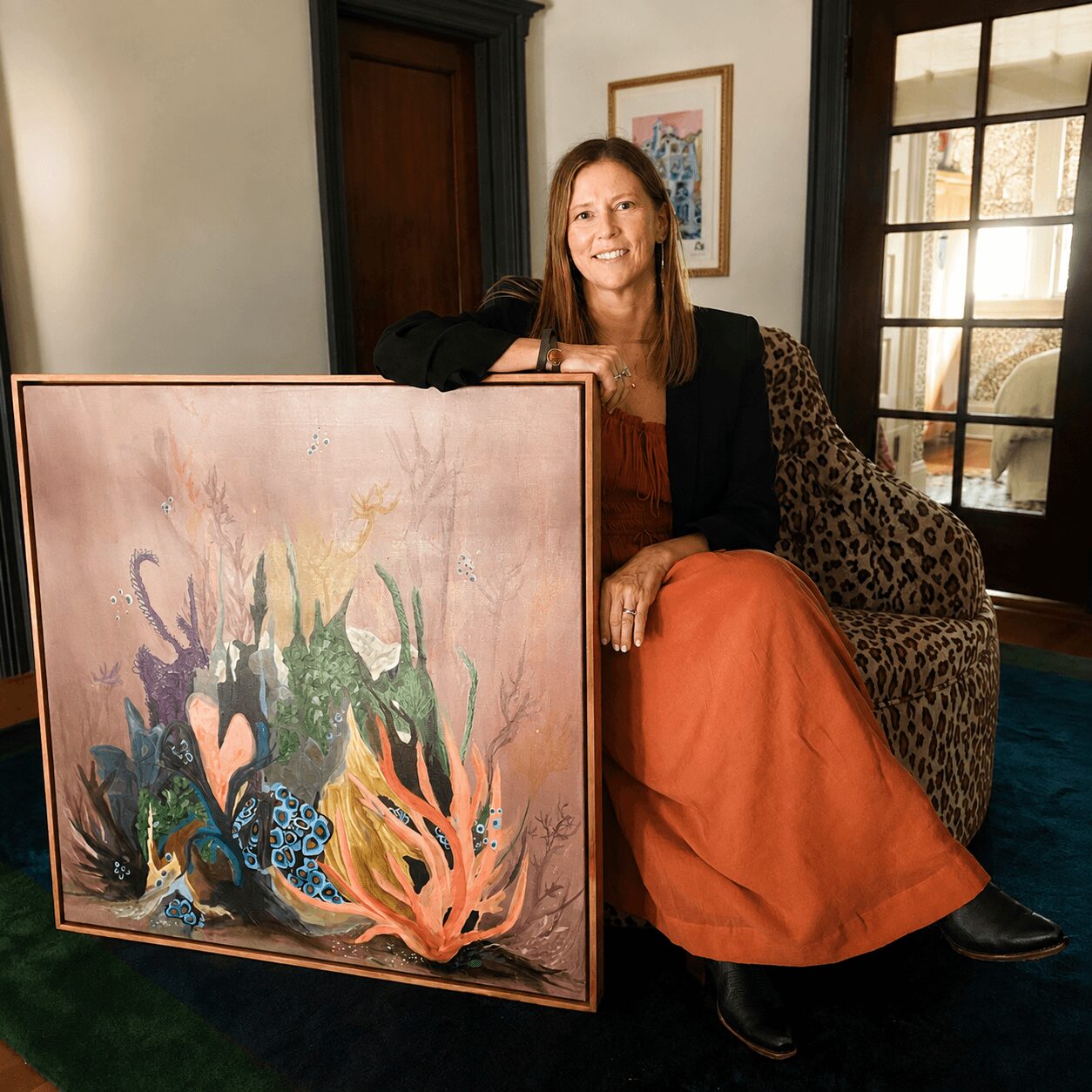 Woman sitting on a leopard-print chair, smiling, next to a large colorful painting in a room with windows and artwork.