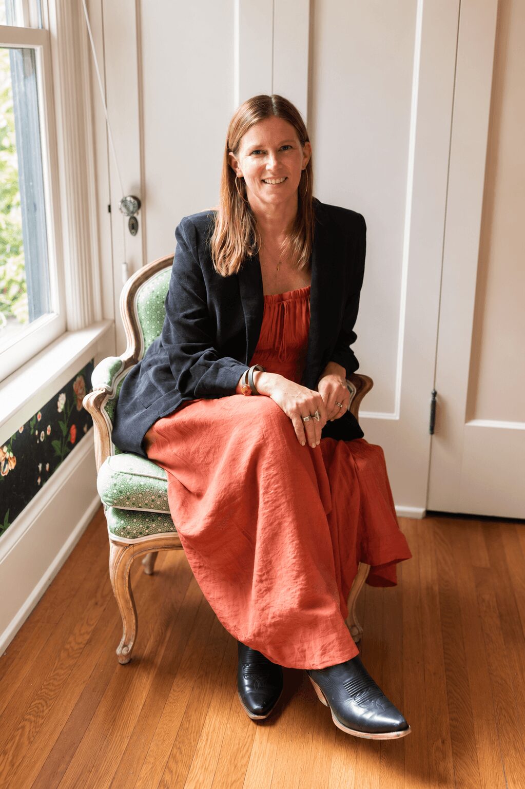 Woman sitting on a vintage chair near a window, smiling, wearing a black blazer and a long red dress, with wooden flooring.