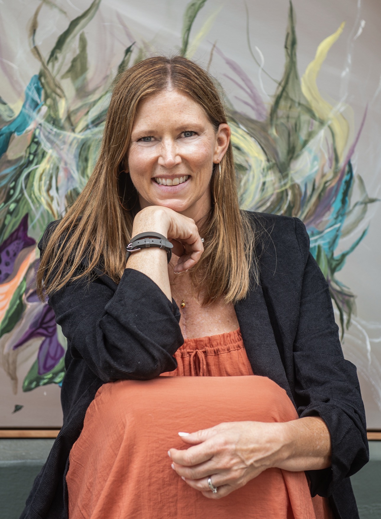 Woman with long brown hair smiling, wearing a black blazer and orange top, sitting in front of colorful abstract background.