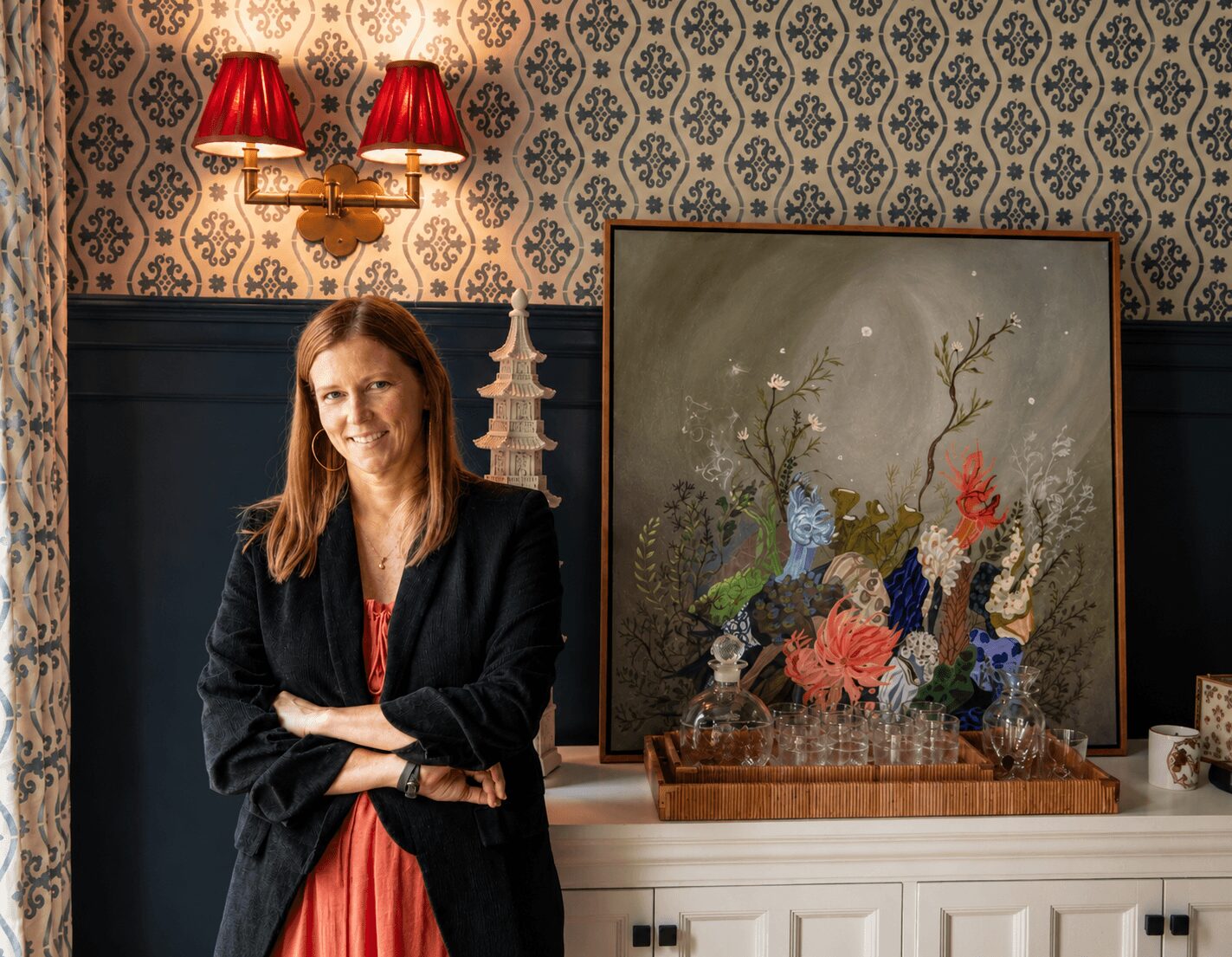 Woman with long hair in black blazer and red dress standing next to a floral painting on a white cabinet, patterned wallpaper background.