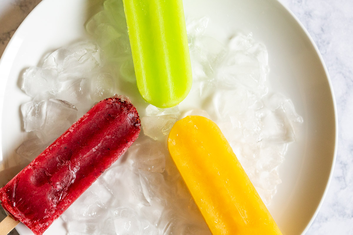 Three colorful popsicles on ice in a white bowl, with green, red, and yellow popsicles visible.