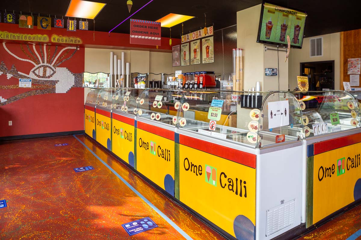 Ice cream shop with yellow and red counters, ice cream displays, and a menu on the wall, with a TV screen overhead.