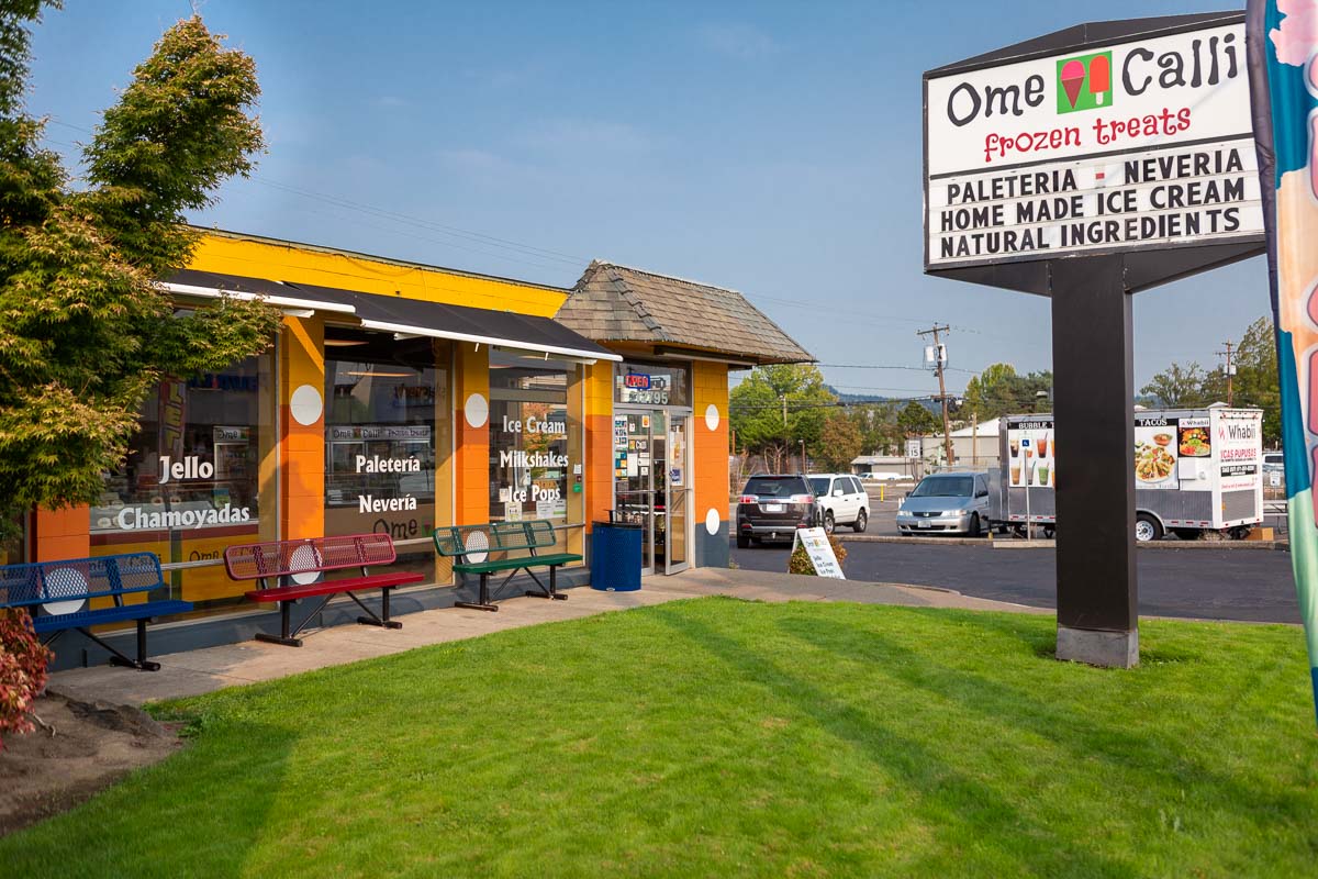 Ice cream shop with yellow awning, benches, and a sign advertising flavors and ingredients, on a sunny day with green grass.