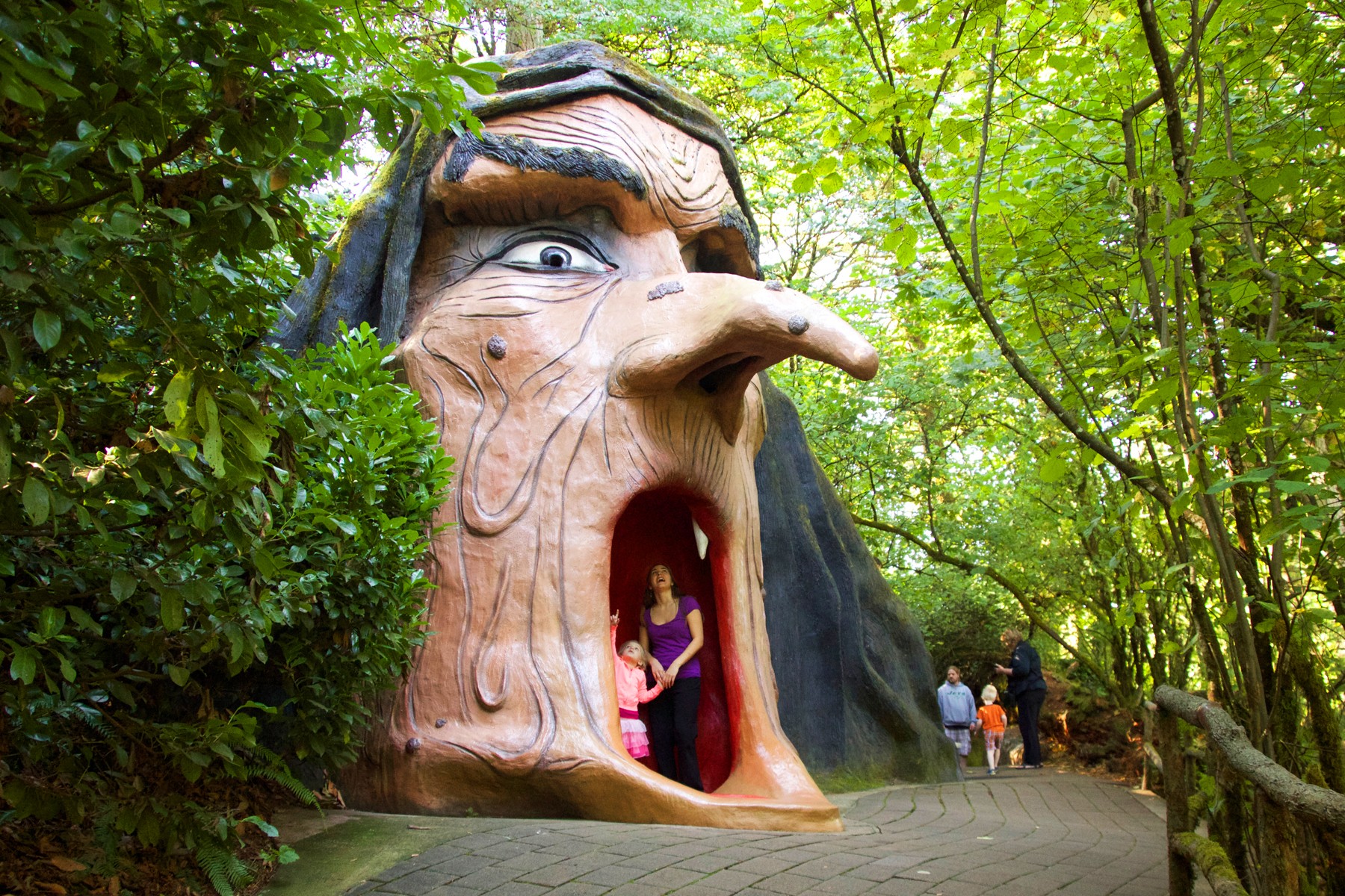 A large witch's head sculpture with an open mouth, surrounded by green trees, with people nearby on a path.