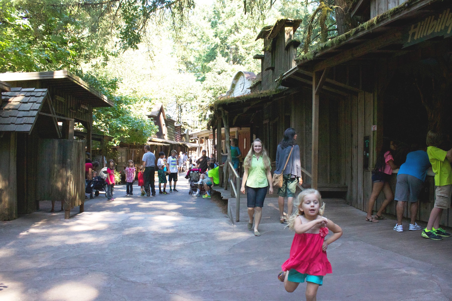 People walking and standing outdoors near wooden structures in a park with trees and sunlight.