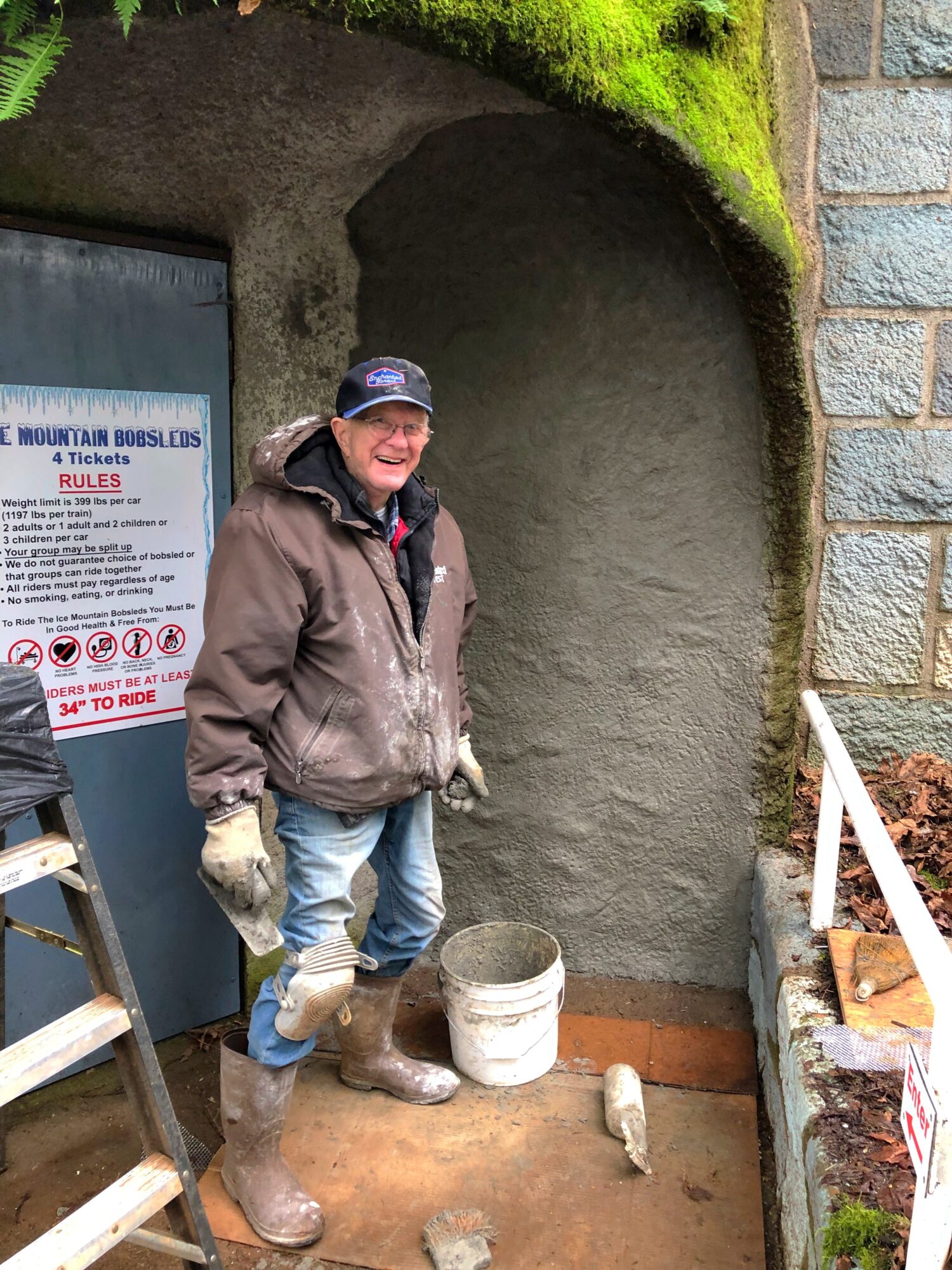 Man in a brown jacket and cap standing next to a partially built wall with a bucket and tools nearby.