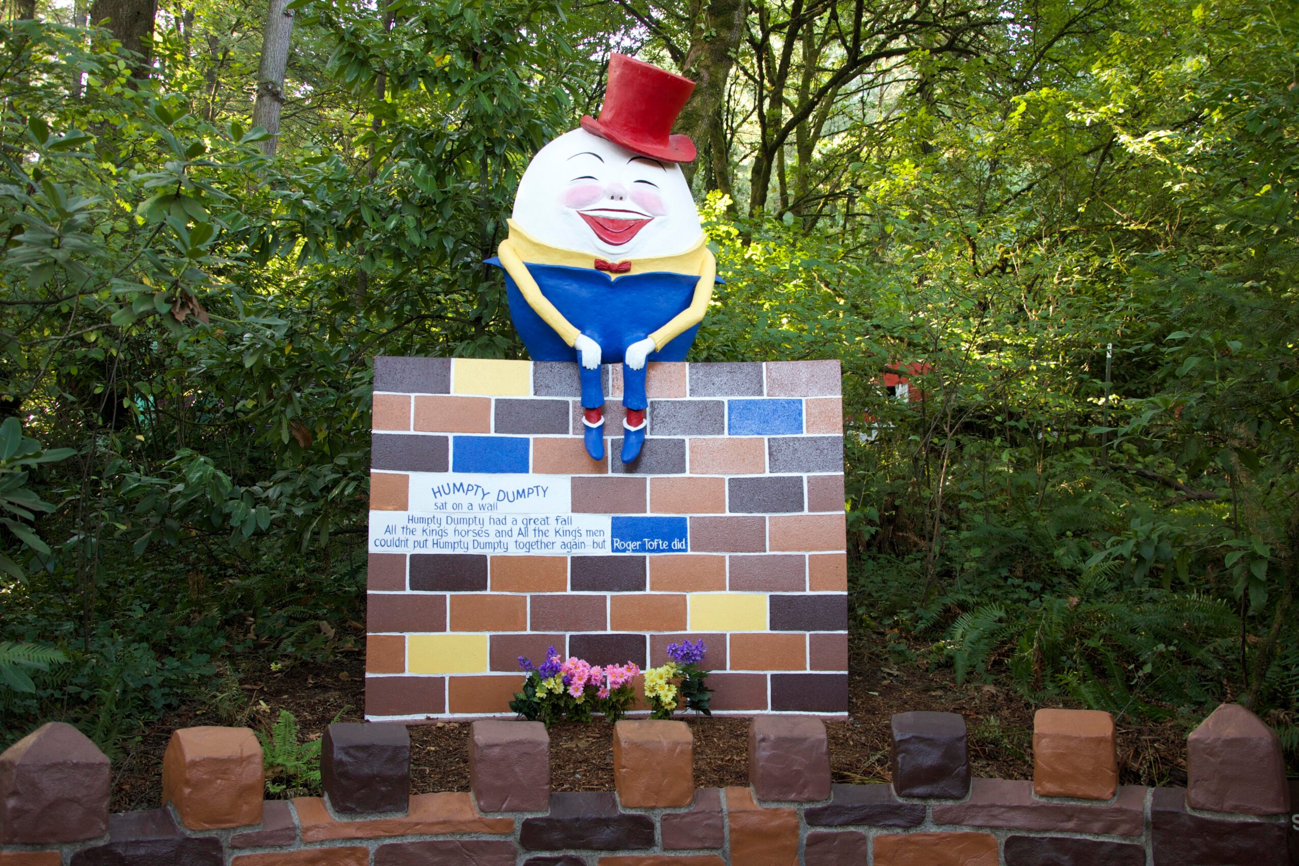 Colorful clown figure with a red hat sitting on a brick wall, surrounded by green trees and bushes, with flowers below.