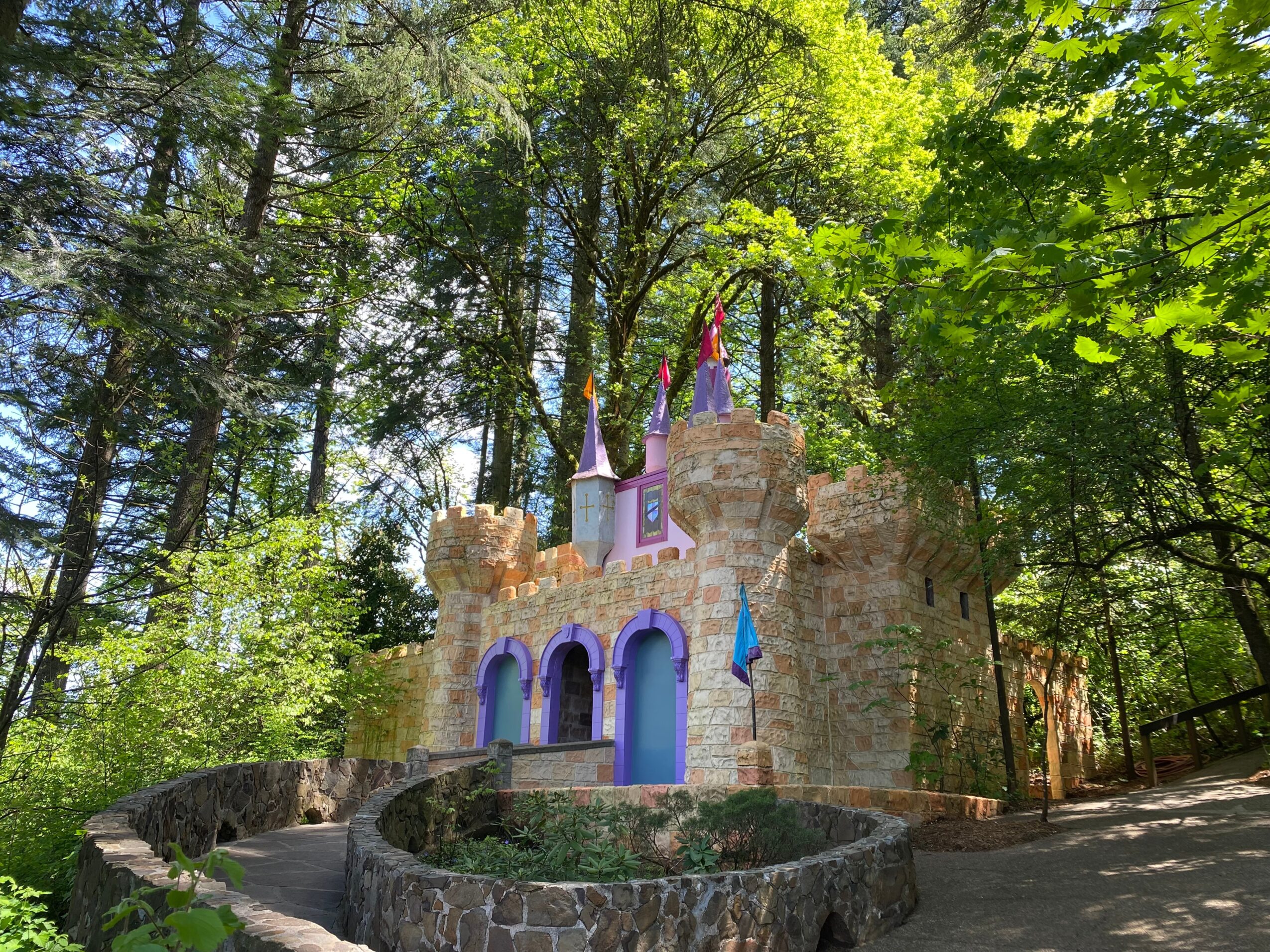 Small colorful castle with purple and pink accents surrounded by green trees and a pathway.