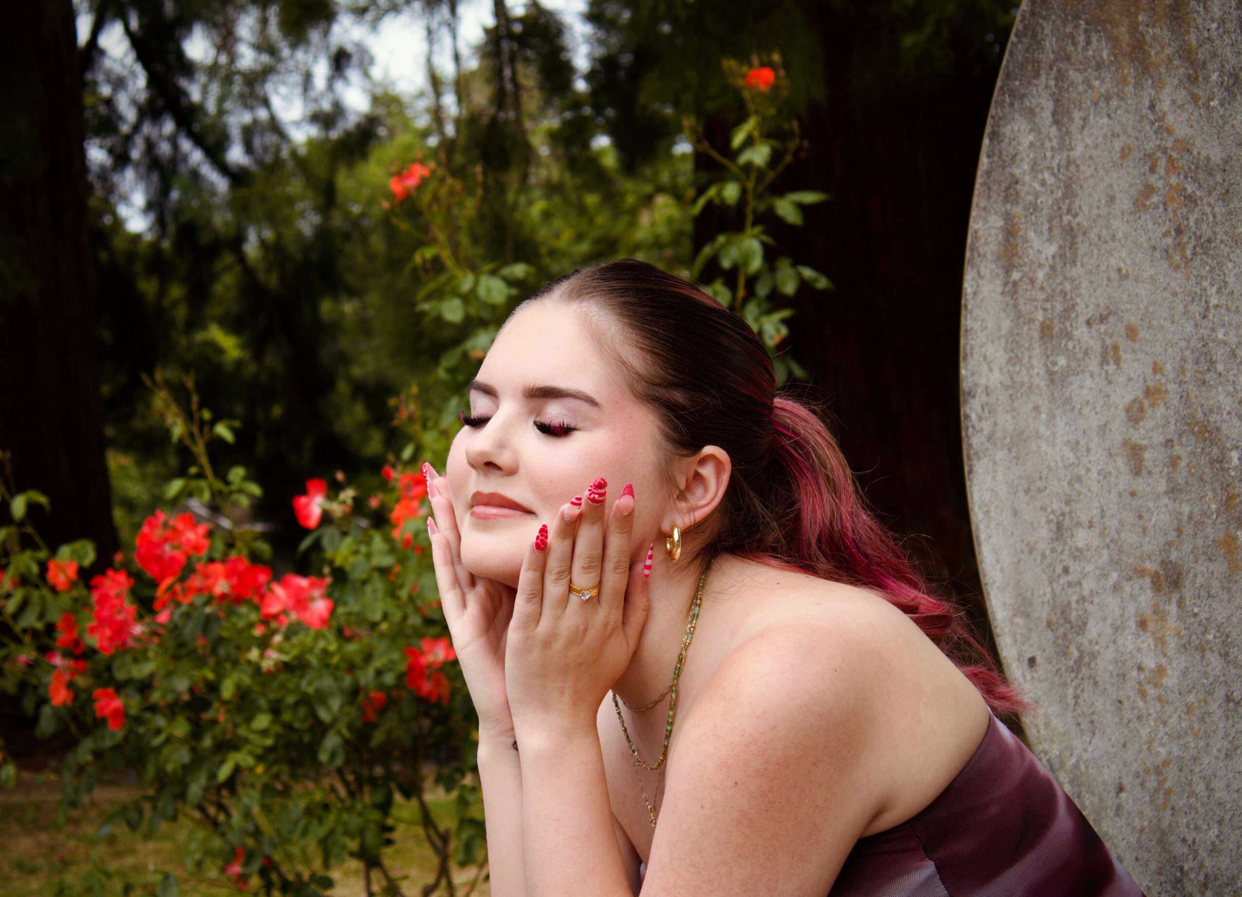 Young woman with closed eyes and hands on face, surrounded by flowers and greenery.
