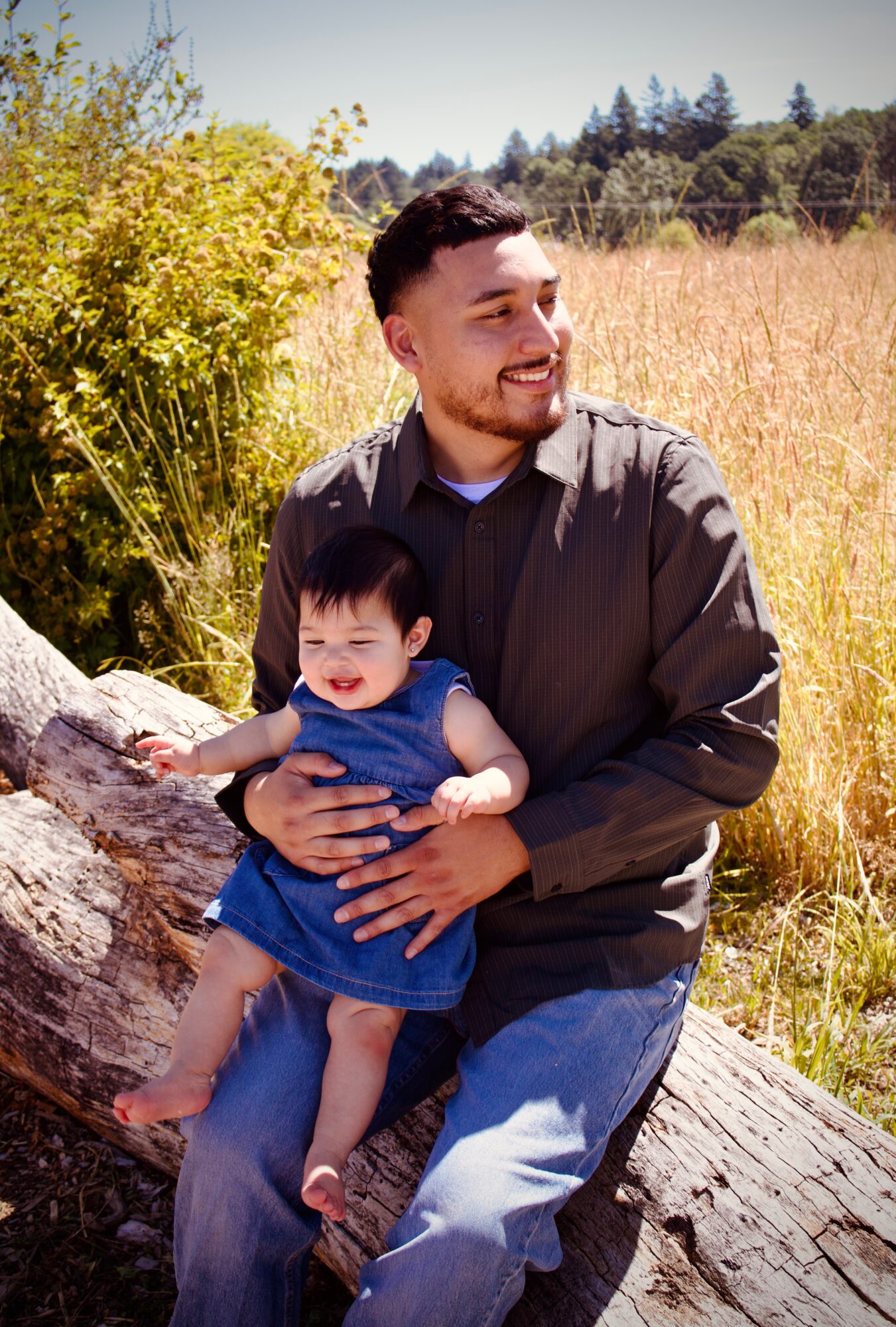 Man sitting on a log holding a smiling young girl outdoors in a field with trees and blue sky.