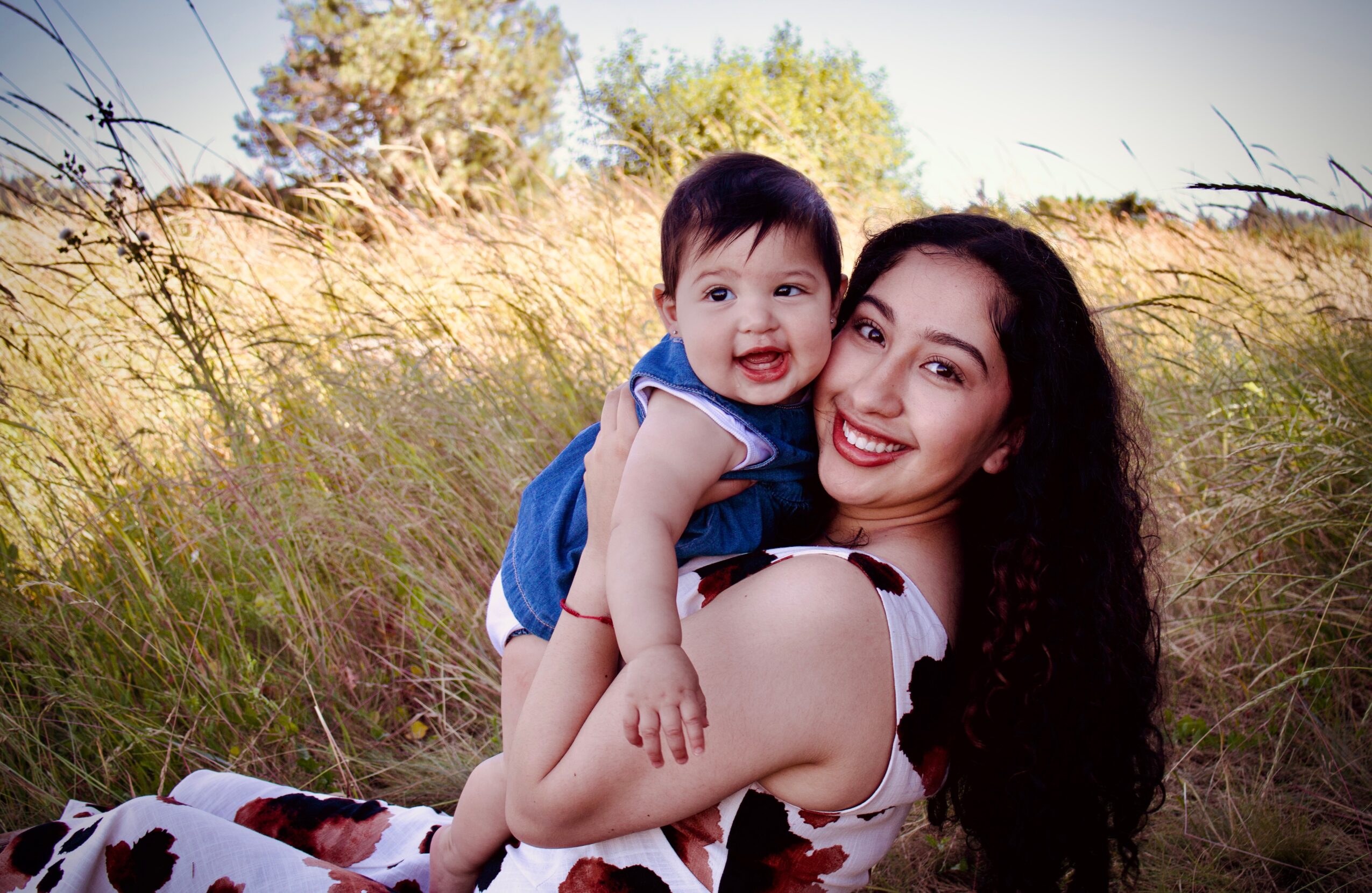 Woman holding smiling child outdoors in a grassy field with trees in background, sunny day.