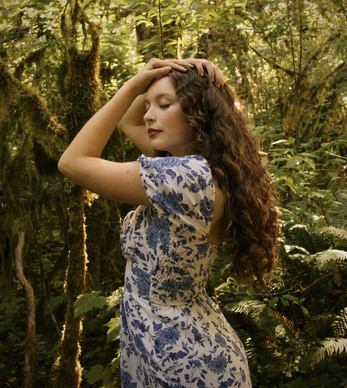 Young woman with long curly hair in a forest, eyes closed, hand on her head, wearing a blue and white patterned dress.