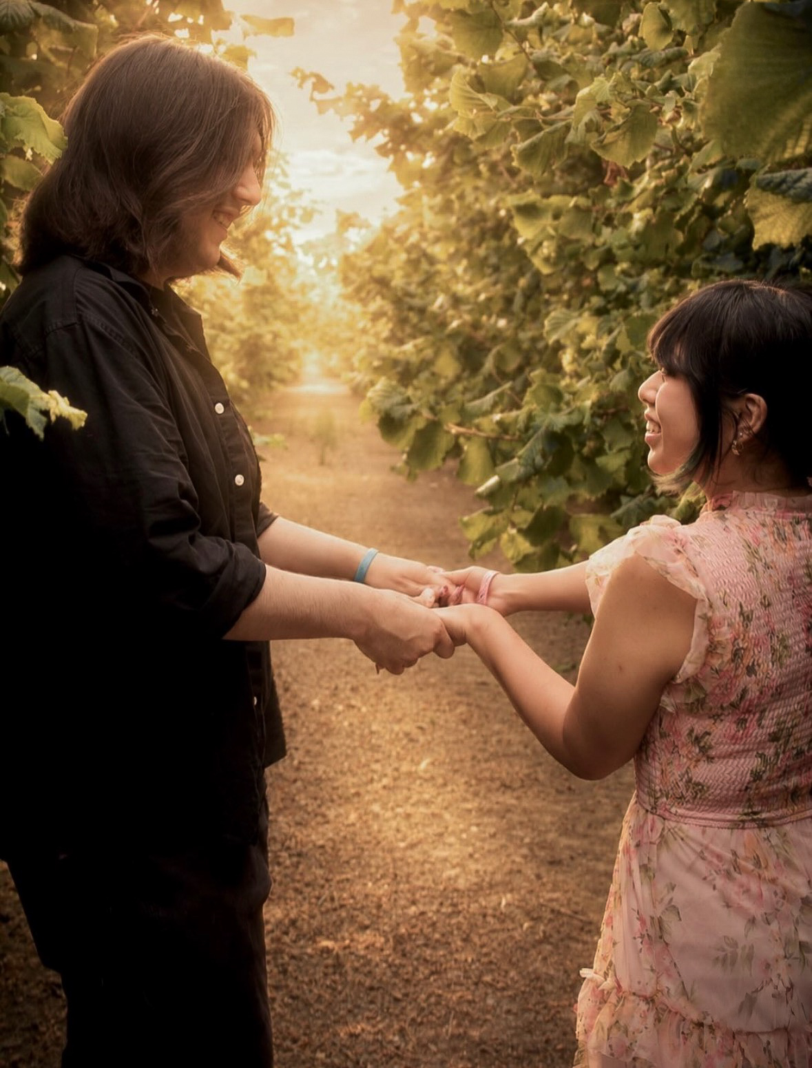 Two women holding hands and smiling in a sunlit outdoor setting with trees and a dirt path.