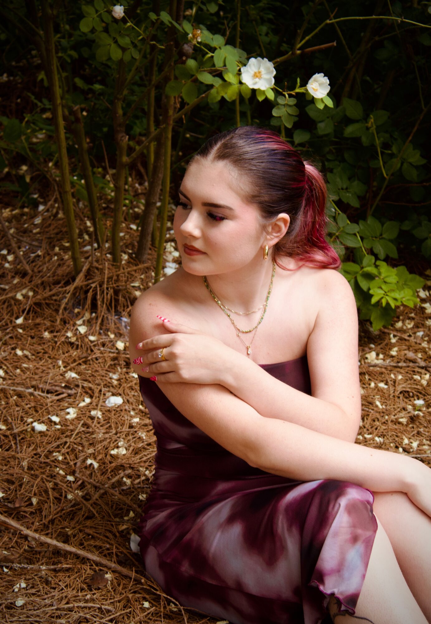 Young woman with red hair in a strapless dress sitting outdoors among greenery and white flowers, looking down thoughtfully.