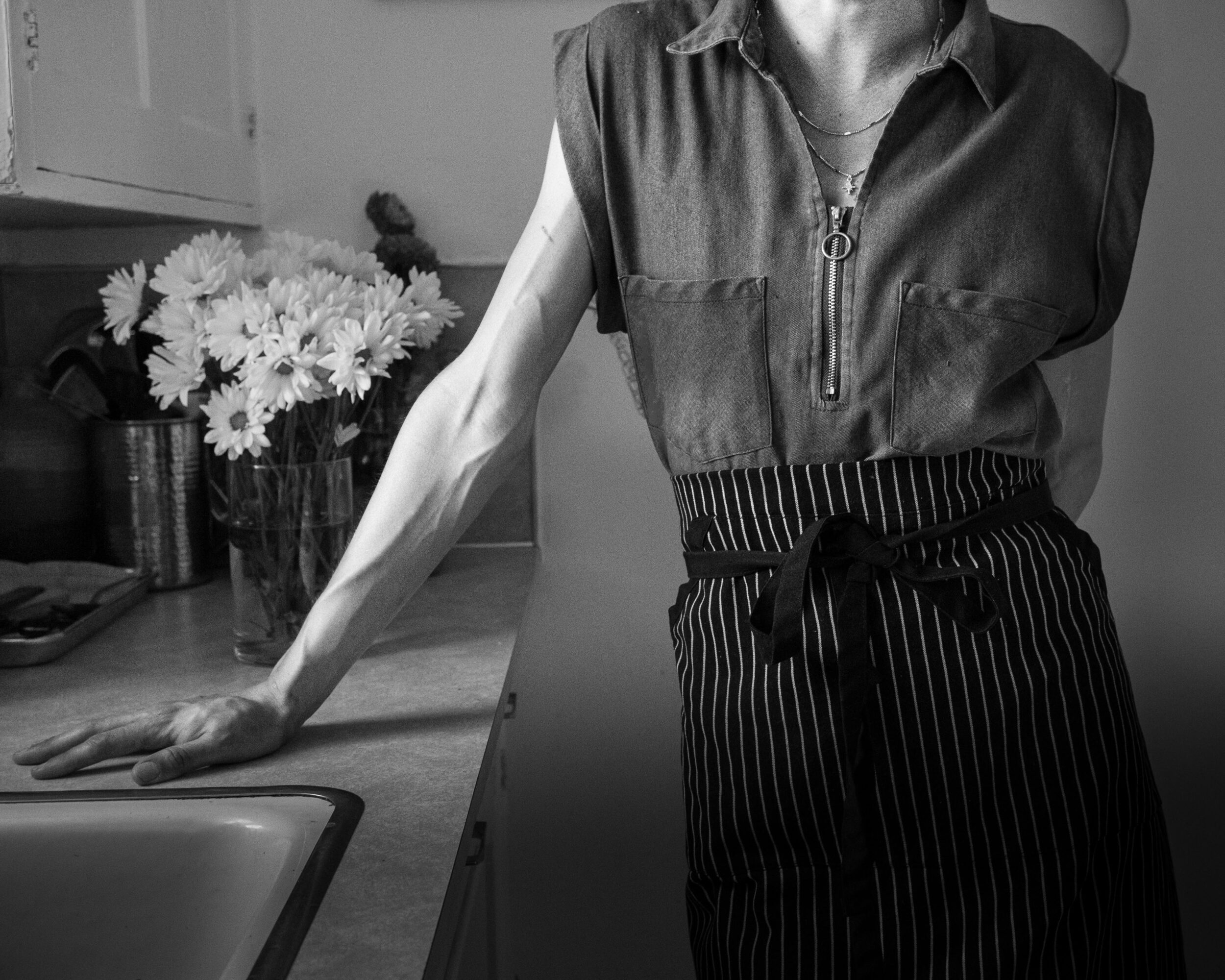 Person leaning on kitchen counter with flowers in background, wearing a sleeveless top and striped apron, in black and white.