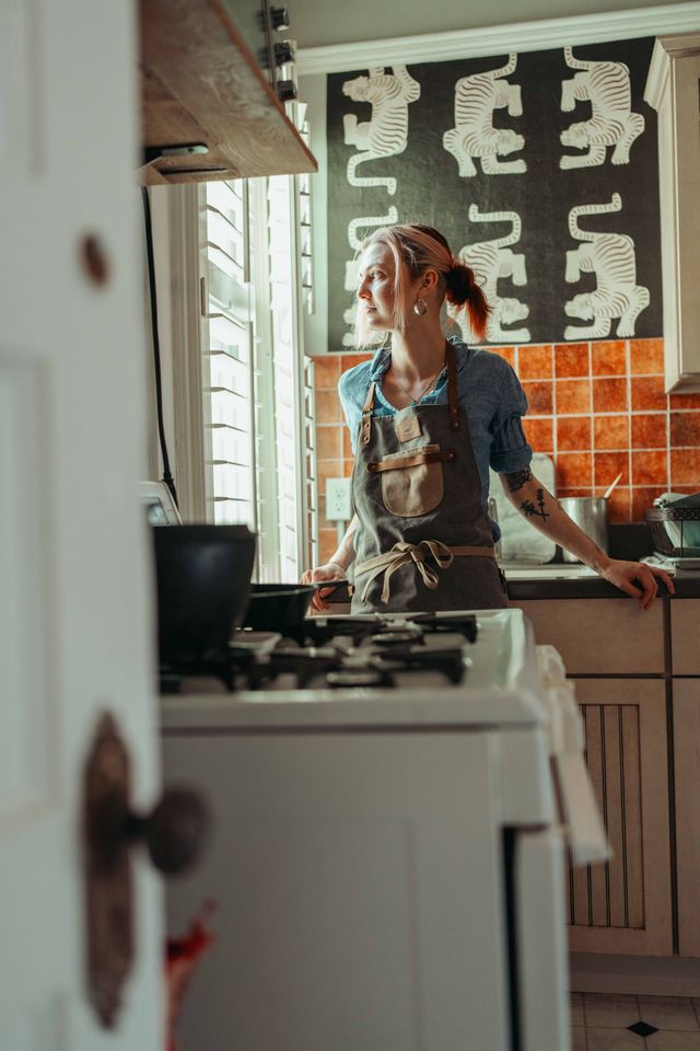 Woman in kitchen looking out window, wearing apron, with patterned black and white curtain, orange tiled wall, and stove in foreground.