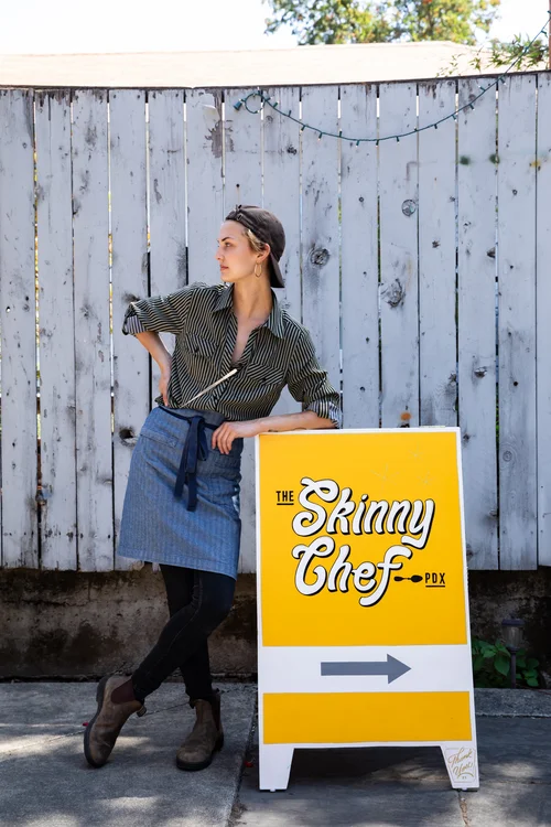 Woman standing next to a sign for The Skinny Chef, pointing right, outdoors, wooden fence background.