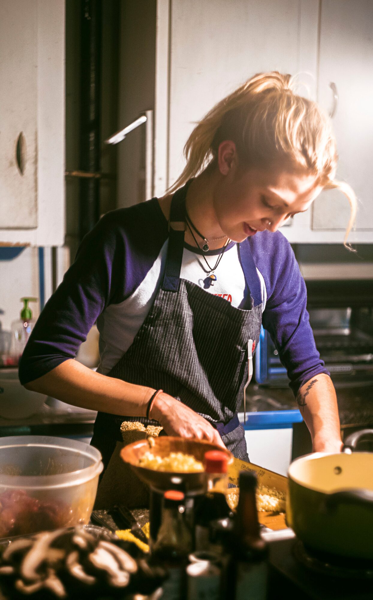 Woman with blonde hair in a ponytail cooking in a kitchen, wearing a black apron and purple shirt, surrounded by cooking utensils and ingredients.
