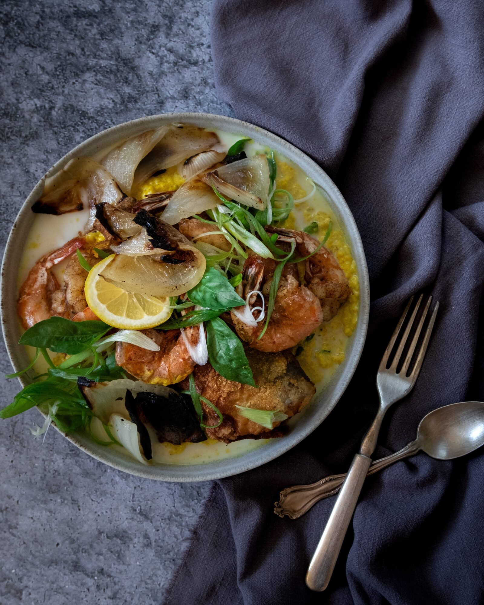Bowl of seafood with shrimp, lemon, vegetables, and herbs on a dark surface with a fork and spoon nearby.