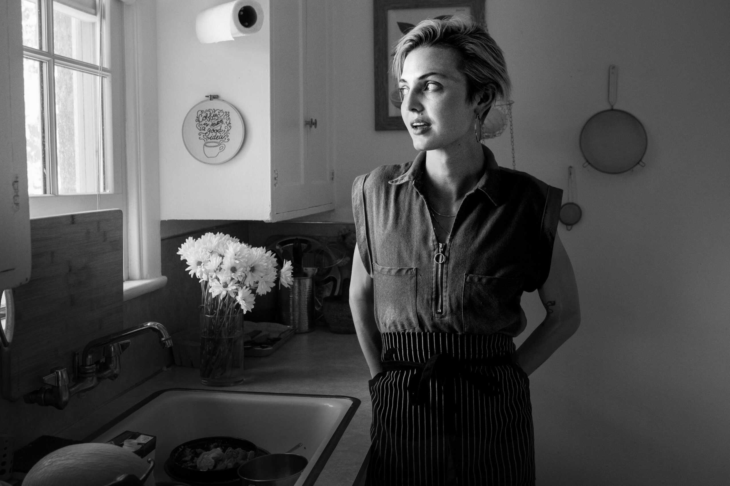 Woman standing in a kitchen with a window, flowers, and kitchen utensils, looking to the side.