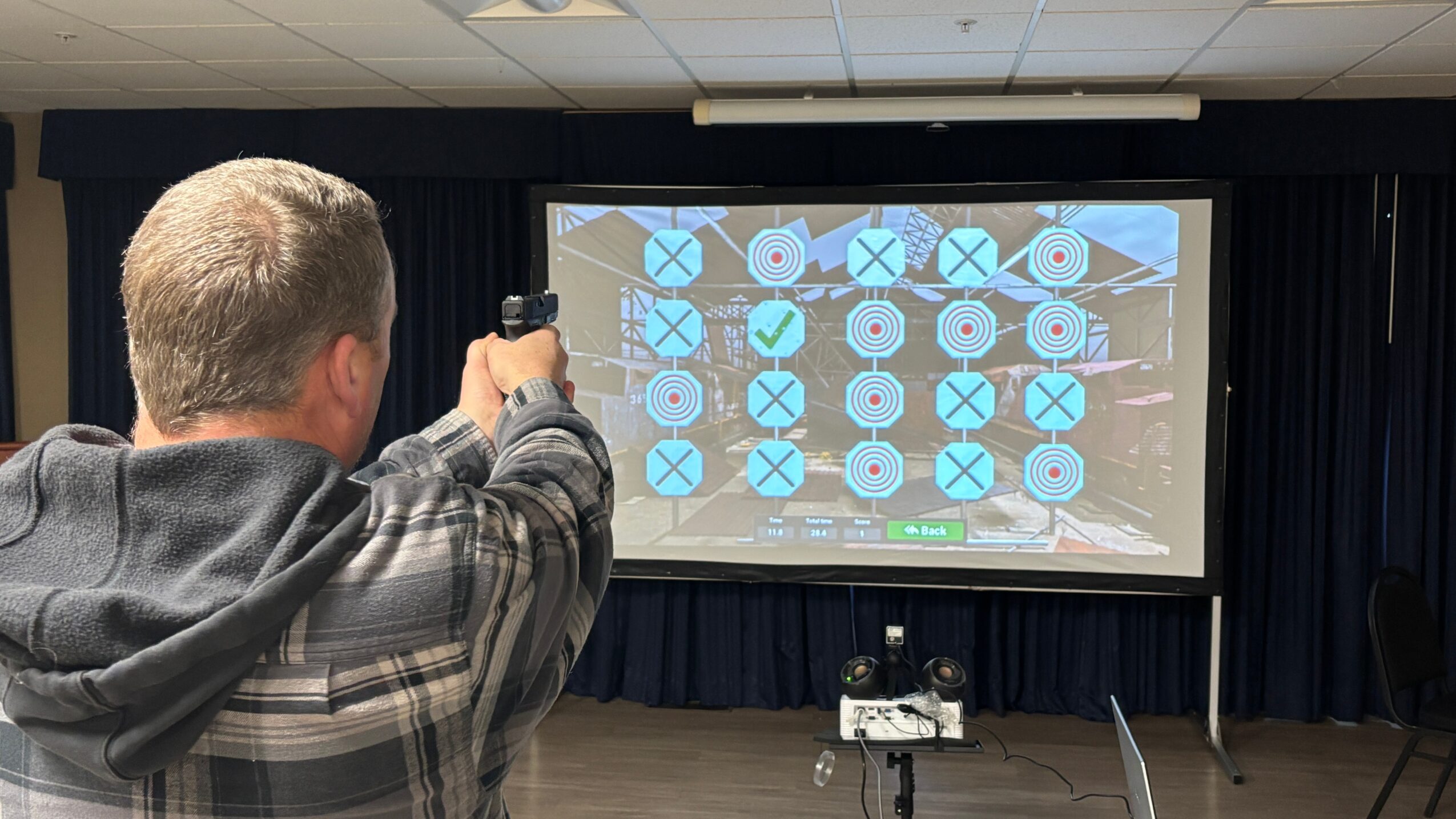 Person aiming a gun at a large screen with a grid of blue circles and checkmarks, in a room with dark curtains.