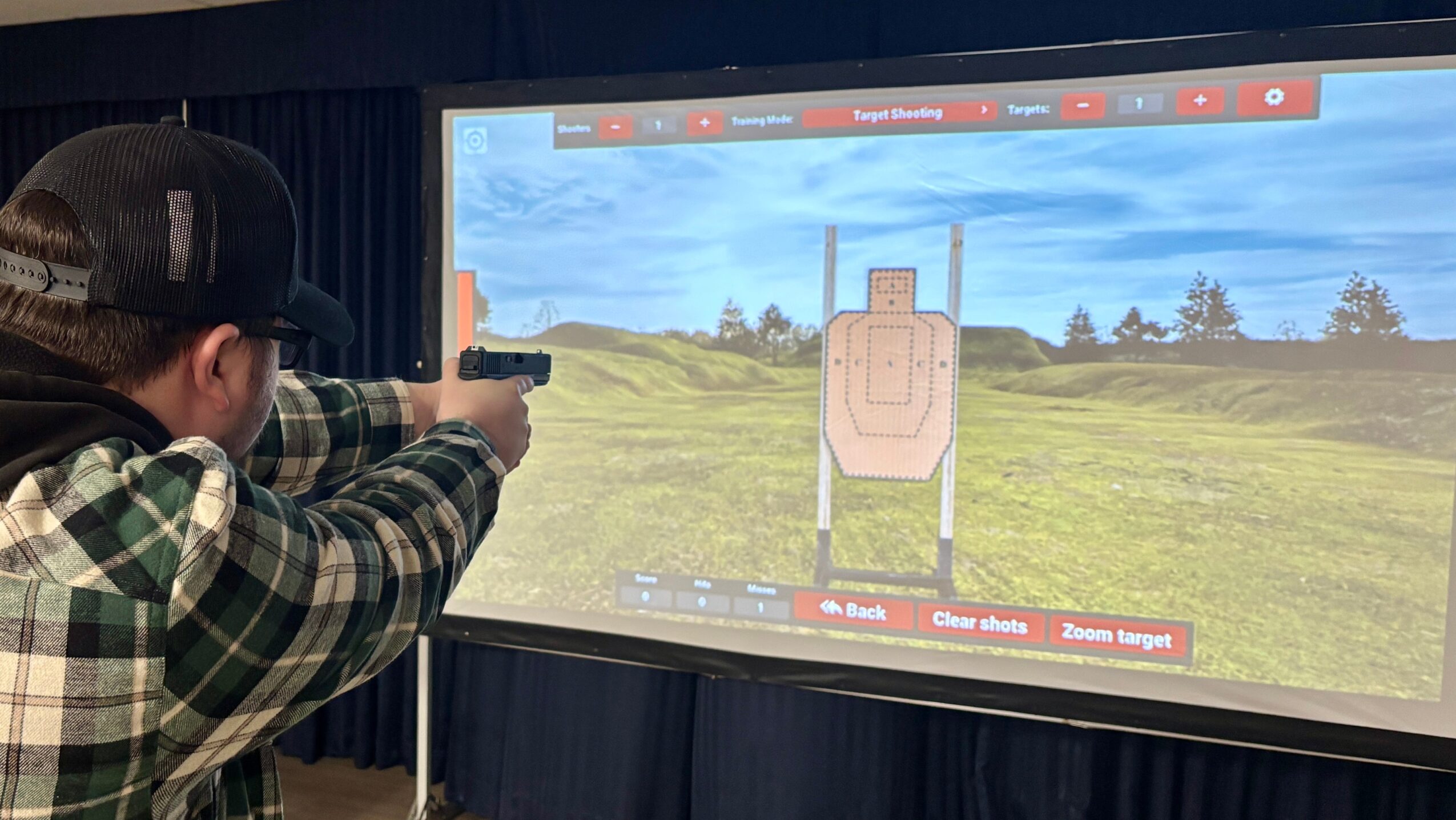 Person aiming a gun at a shooting target on a large screen, with a grassy field and blue sky background.