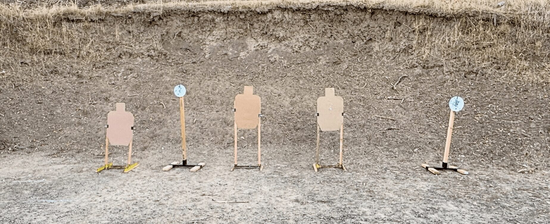 Five wooden targets with blue circles on top are placed on a dirt ground, with a dirt wall behind them.