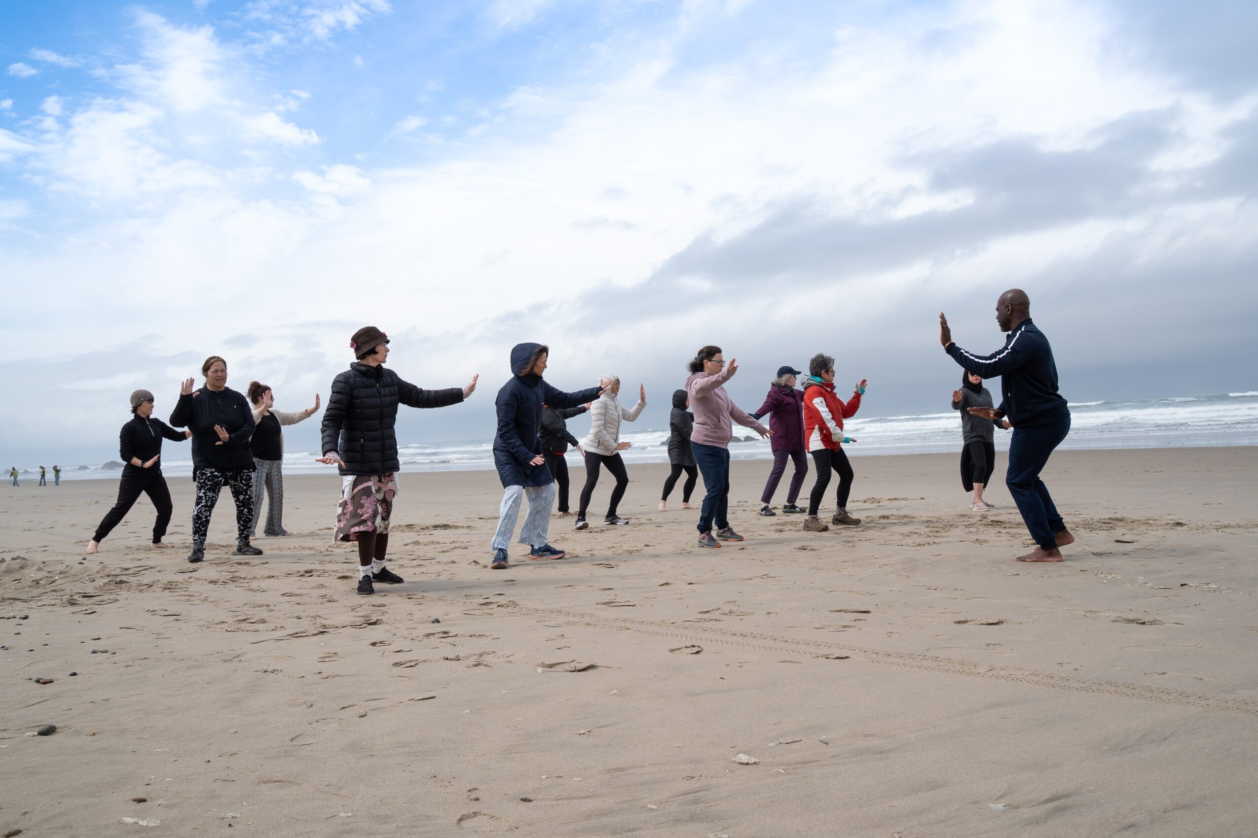 Group of people dancing on a beach with ocean and cloudy sky in background.