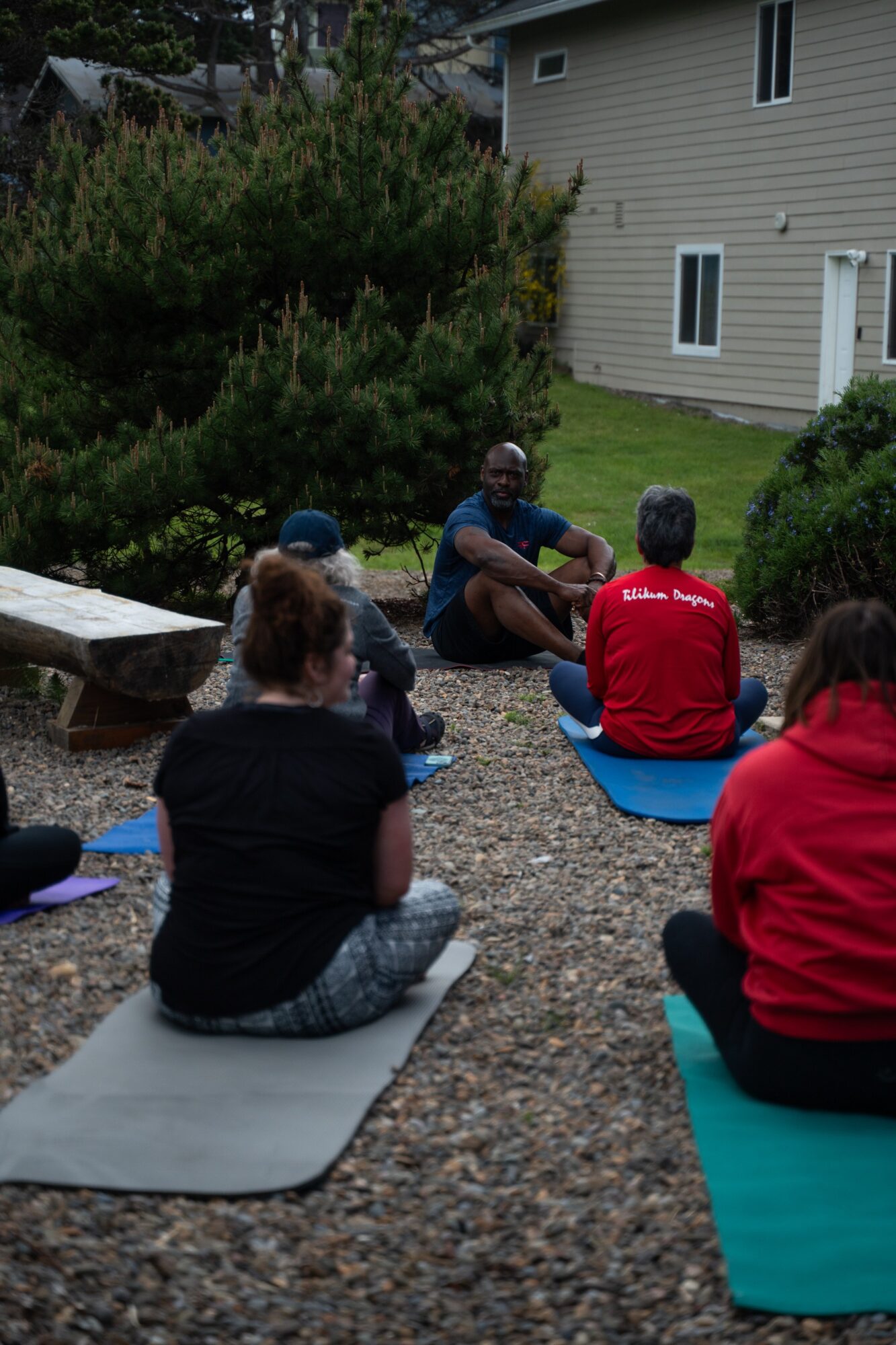 Group of people sitting outdoors on mats and a blue blanket, listening to a person speaking, with a house and greenery in background.