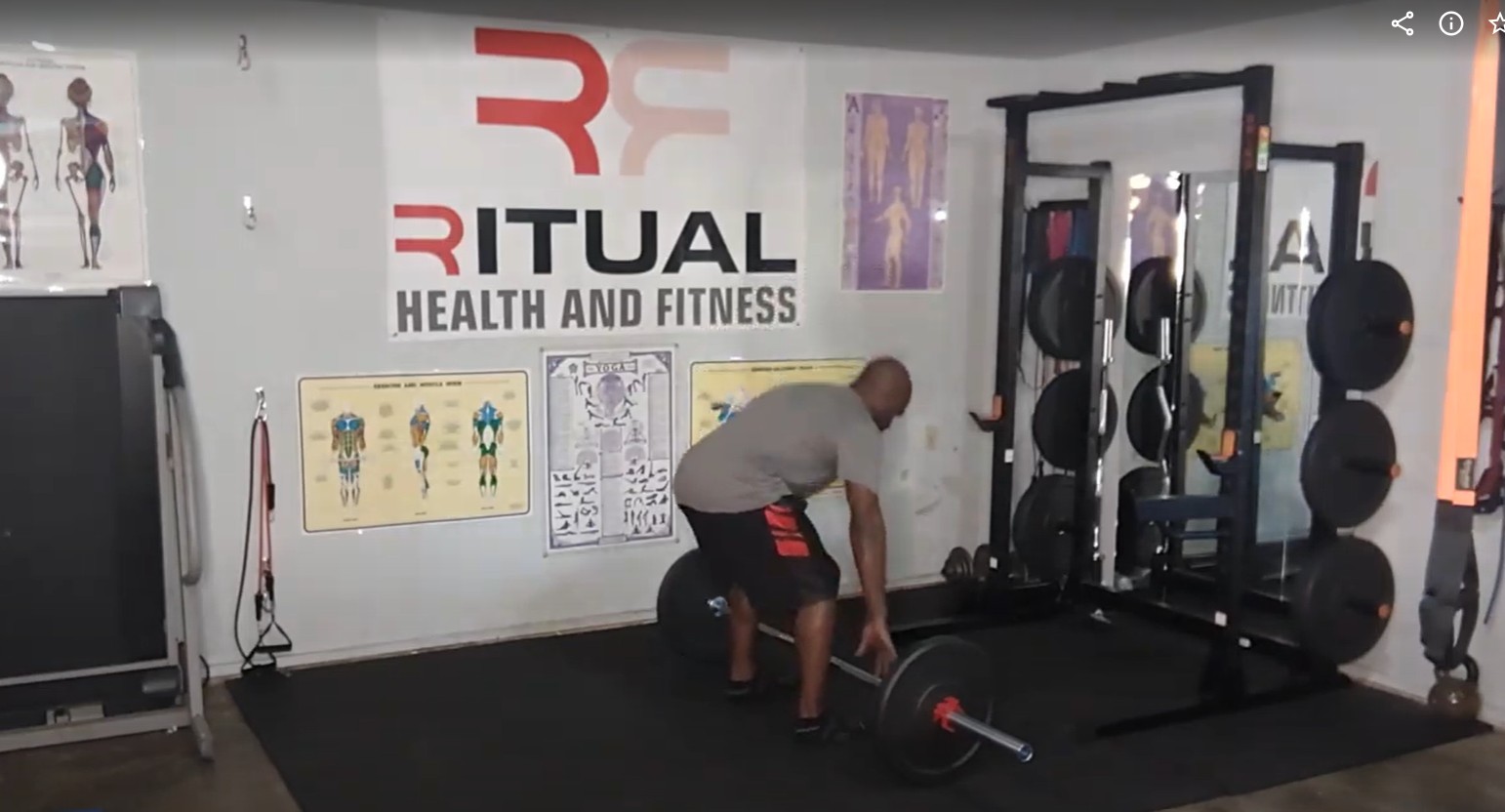 Man lifting a barbell in a gym with fitness posters and equipment, including weights and a squat rack.