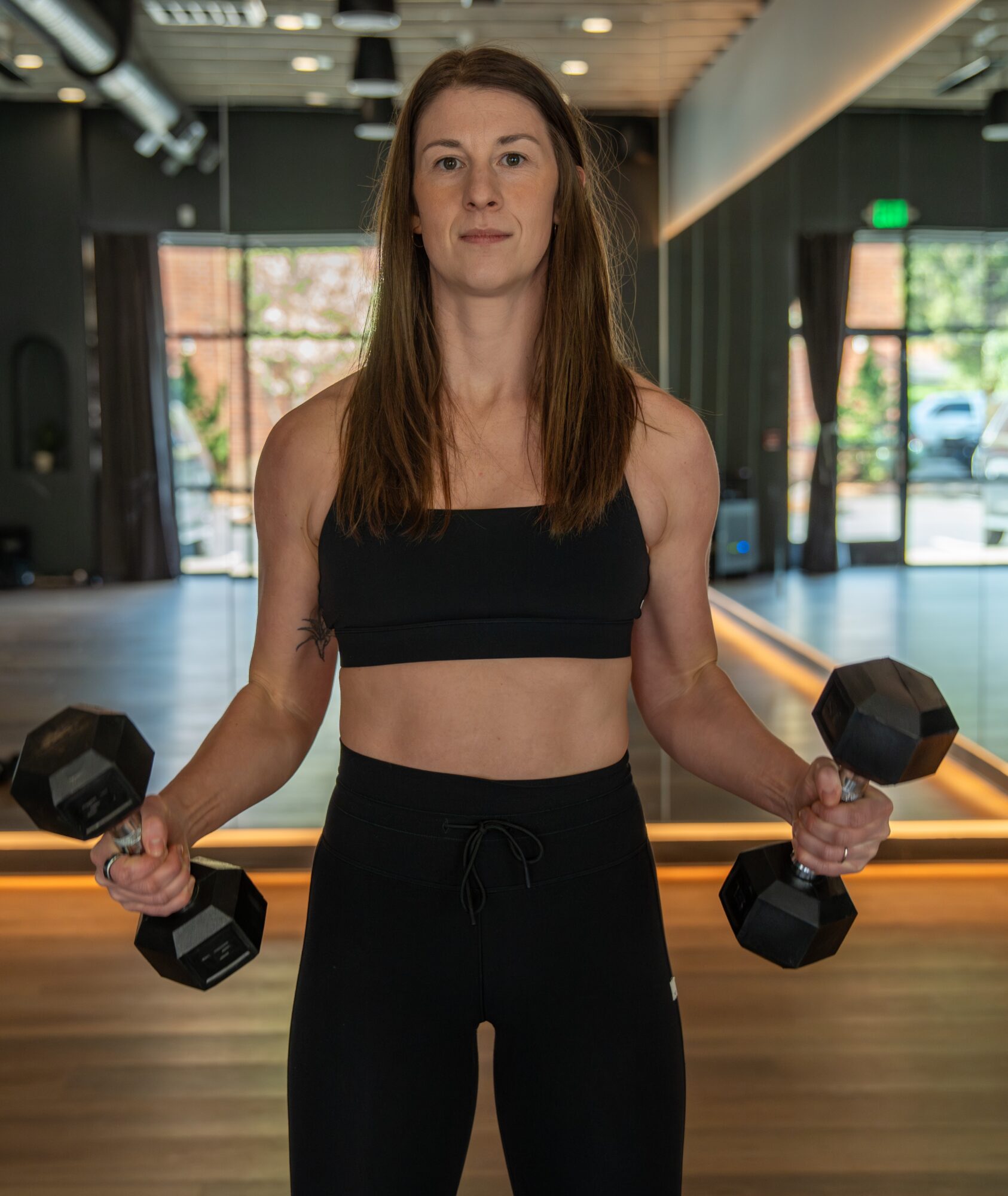 Woman in gym holding dumbbells, wearing black sports bra and pants, with gym interior and windows in background.