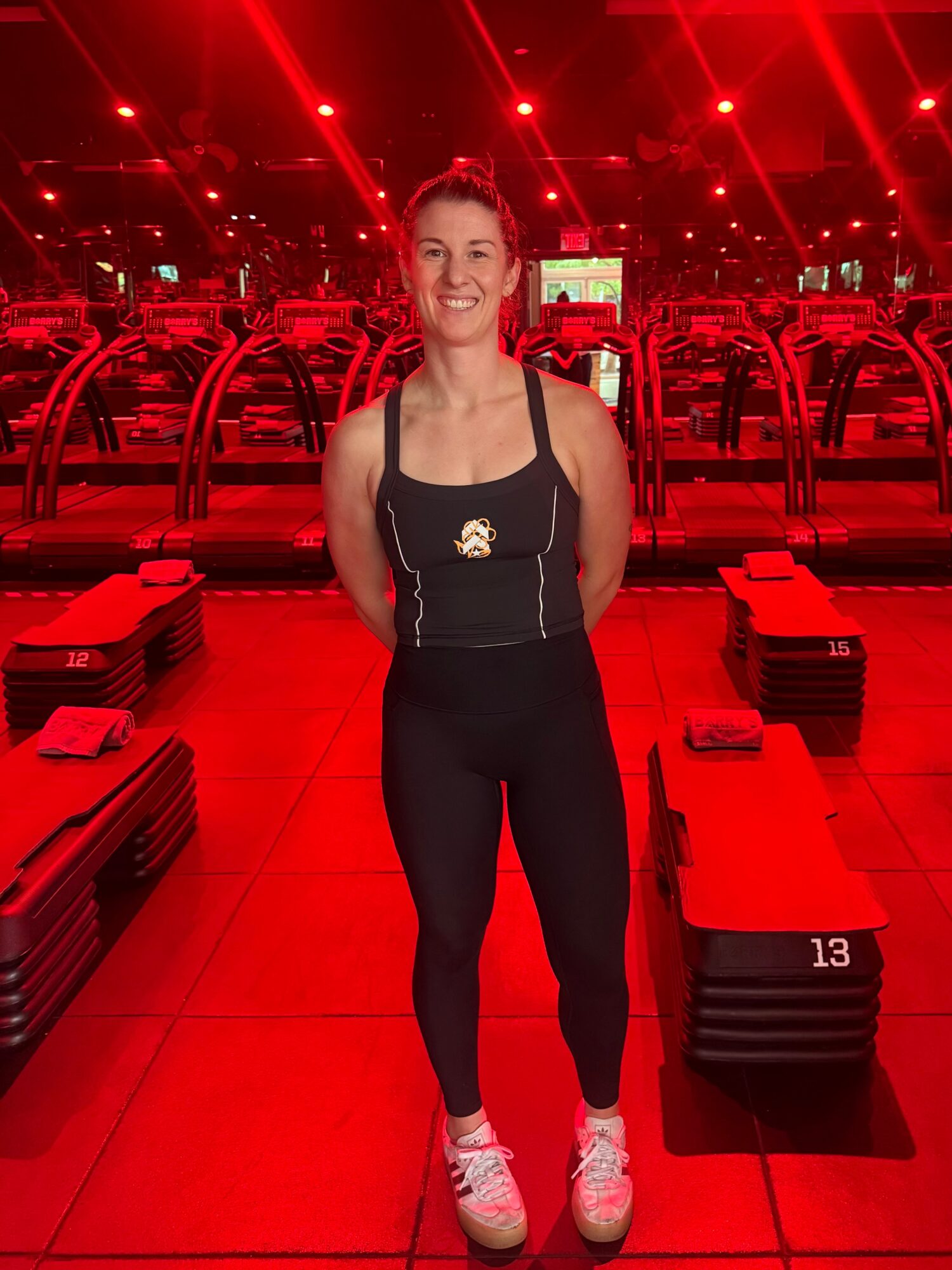 Smiling woman in workout clothes standing in a gym with red lighting and exercise equipment behind her.