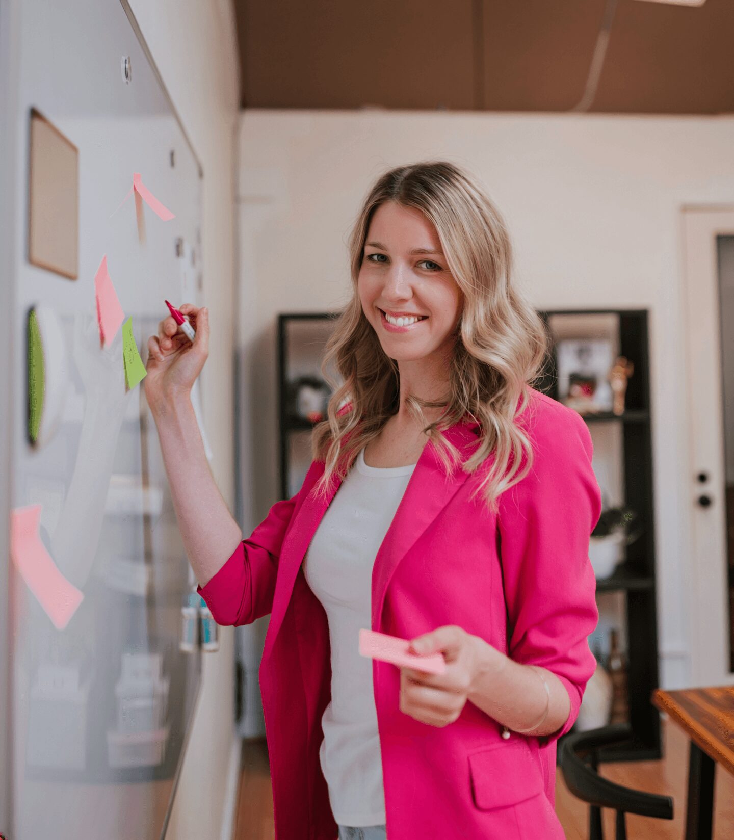 Smiling woman in pink blazer writing on a whiteboard with sticky notes, holding a pink sticky note, in a room with shelves.