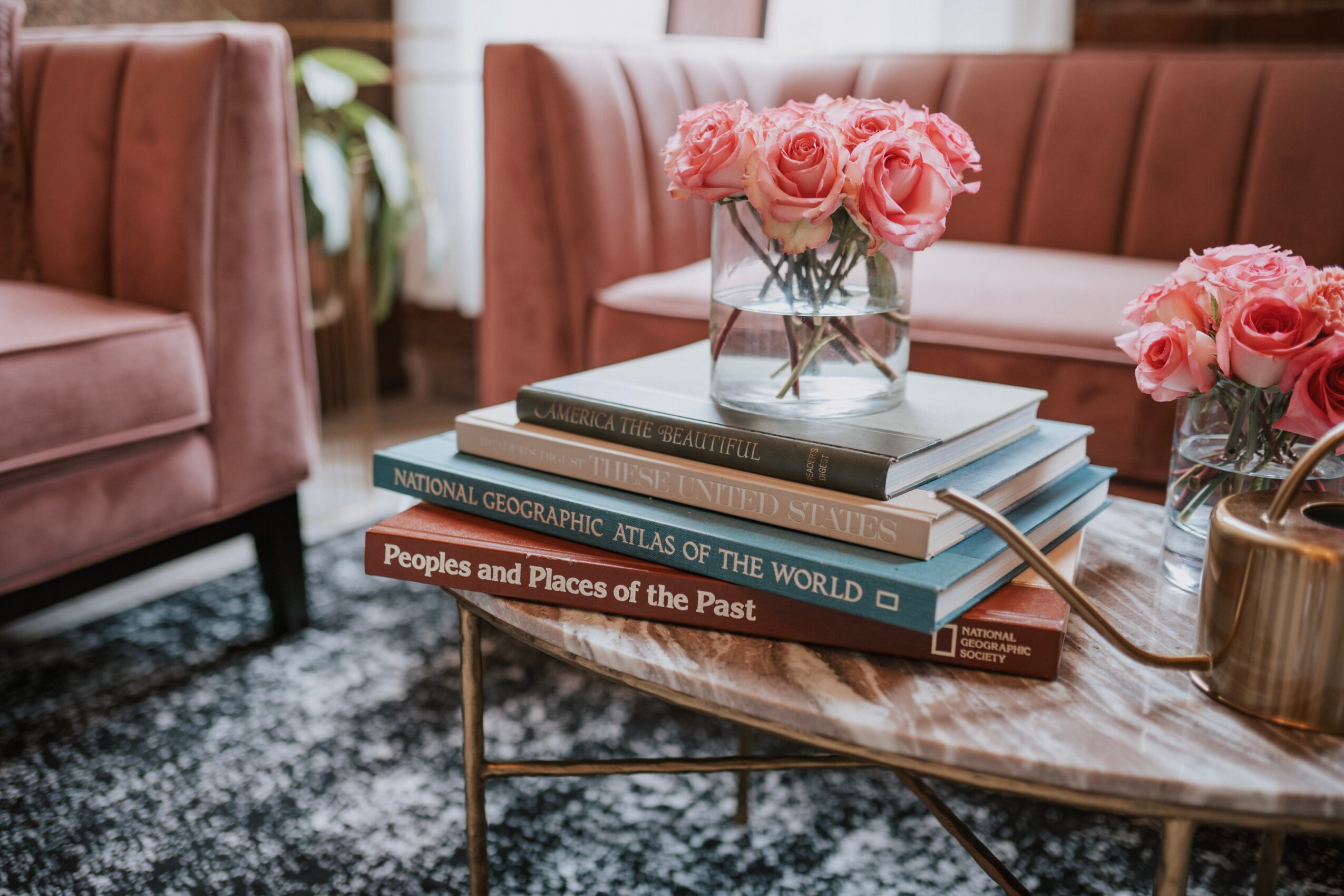 Pink roses in glass vases on a coffee table with stacked books, in a cozy living room setting.