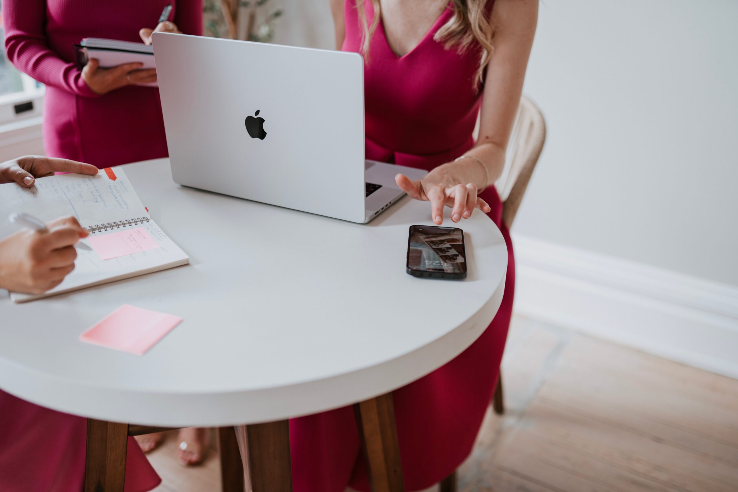 Person in pink dress working on a laptop at a white table with a phone and notepad.