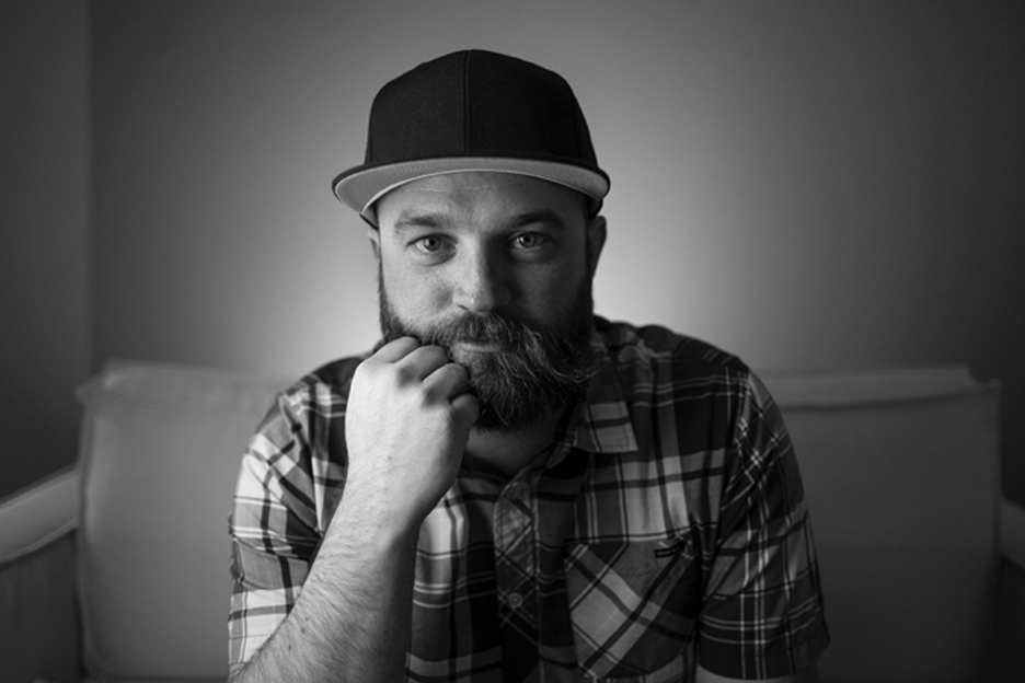 Man with a beard wearing a cap and plaid shirt, resting his chin on his hand, looking at the camera, in black and white.