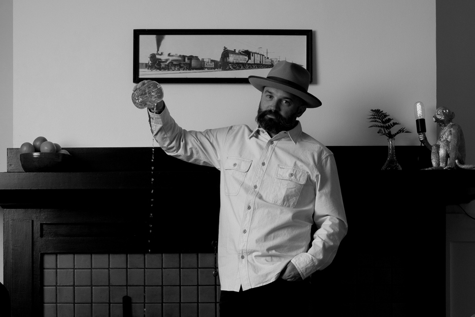 Man with a beard wearing a hat and white shirt, holding a pocket watch, standing indoors with decorative items.