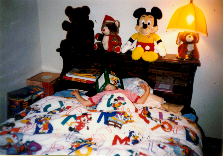 Child sleeping in bed with colorful Mickey Mouse-themed blanket, stuffed Mickey and Minnie Mouse toys on headboard, and a yellow lamp. Word count: 24